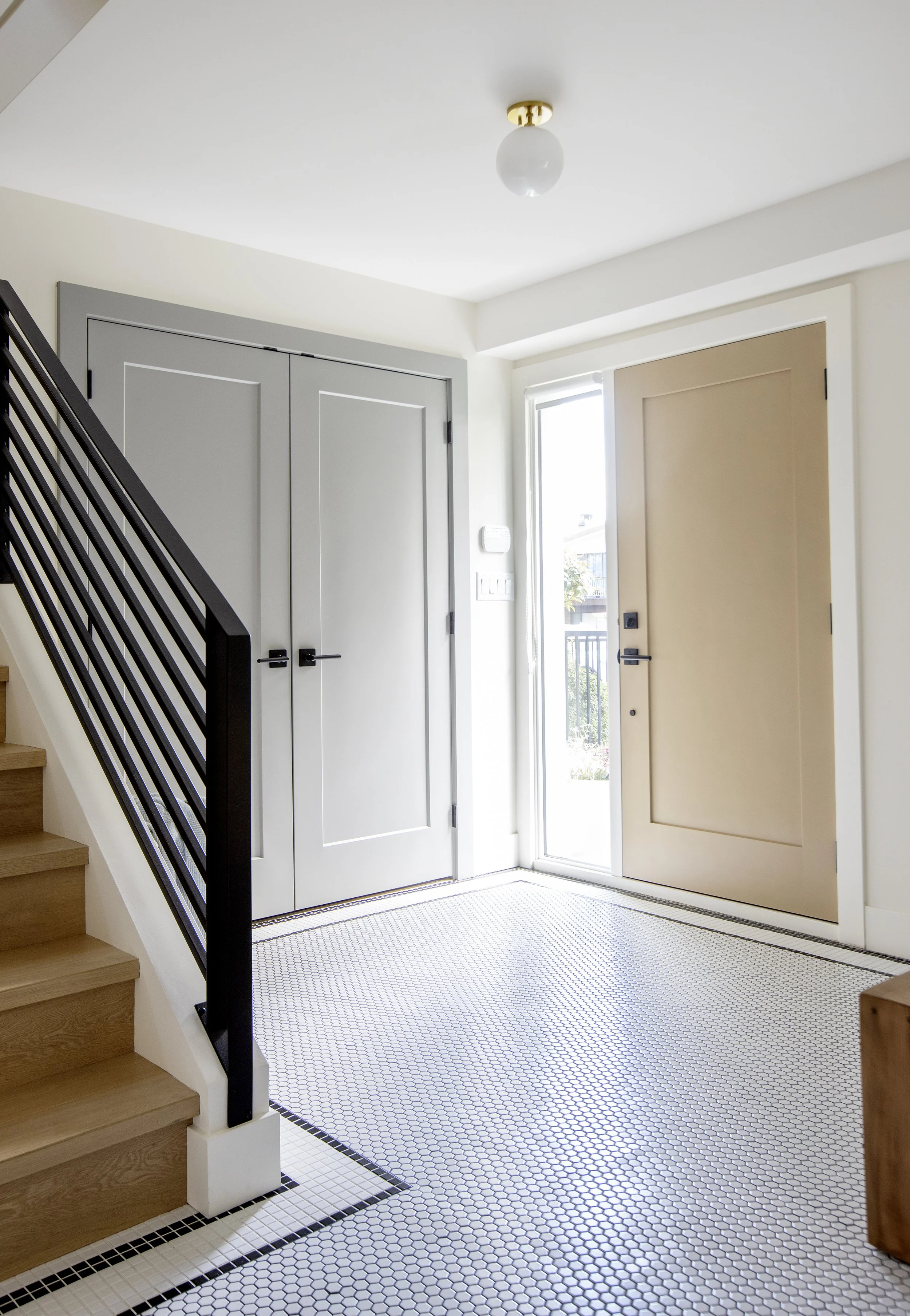 Interior entryway with black and white hexagonal tile floor, staircase with wooden steps and black metal railing, light-colored closet doors, and a glass front door allowing natural light.