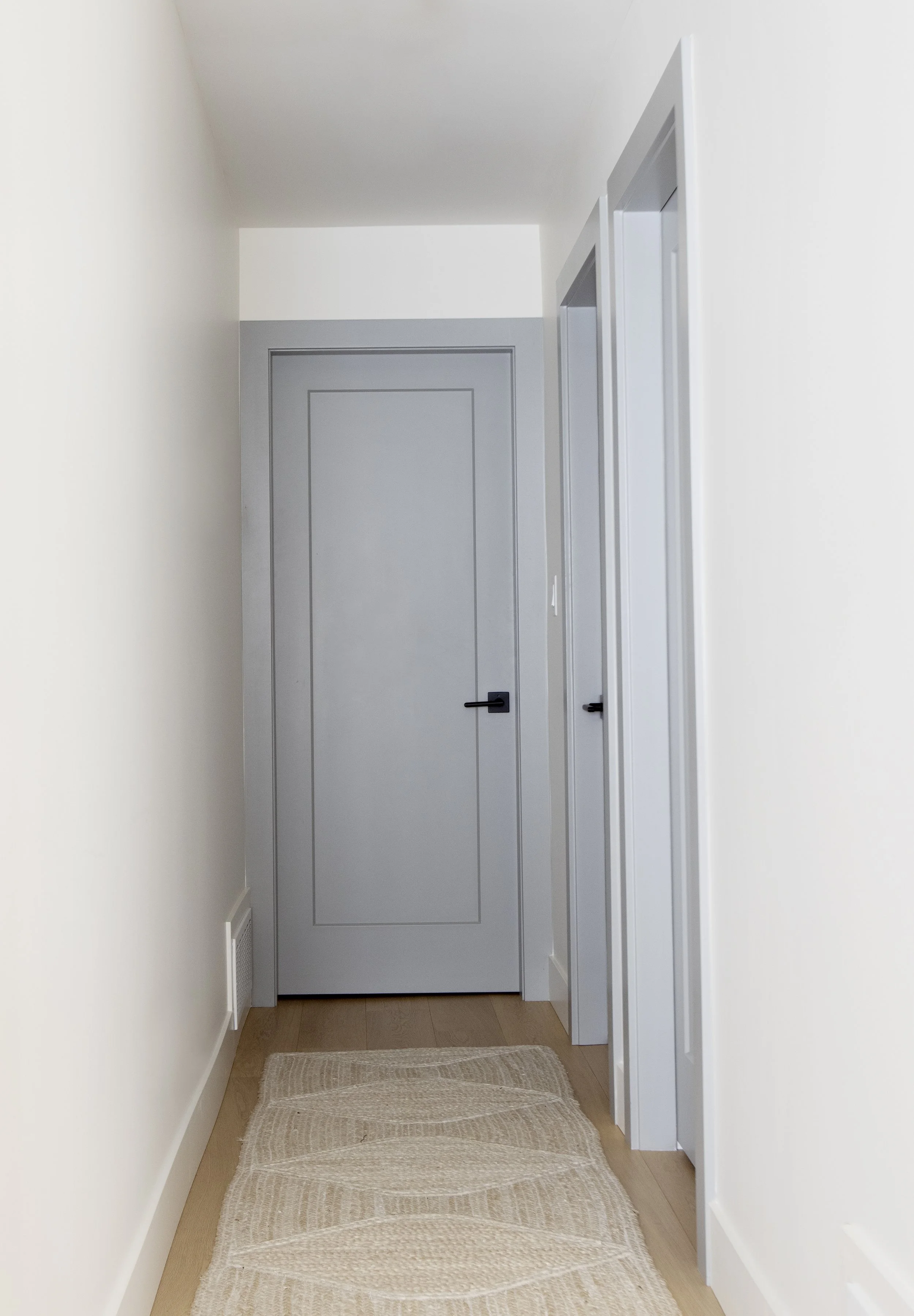 A narrow hallway with white walls, a light-colored wooden floor, a closed gray door at the end, and two white doors on the right side. There is a beige woven rug on the floor near the entrance.