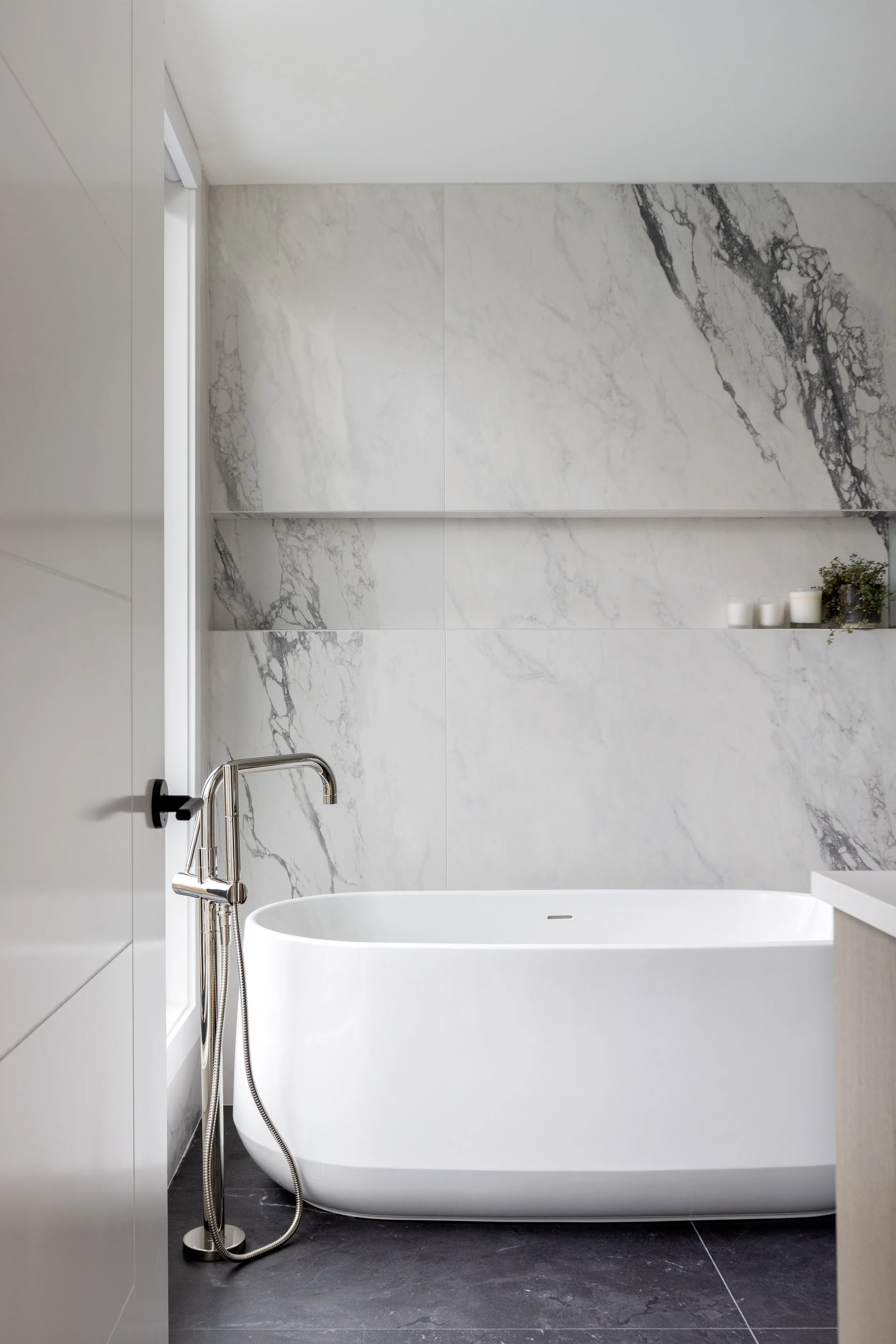 Modern bathroom with a white freestanding bathtub, gray marble wall tiles, black floor tiles, and a chrome floor-mounted bathtub faucet with a handheld showerhead. A small shelf holds candles and a plant.