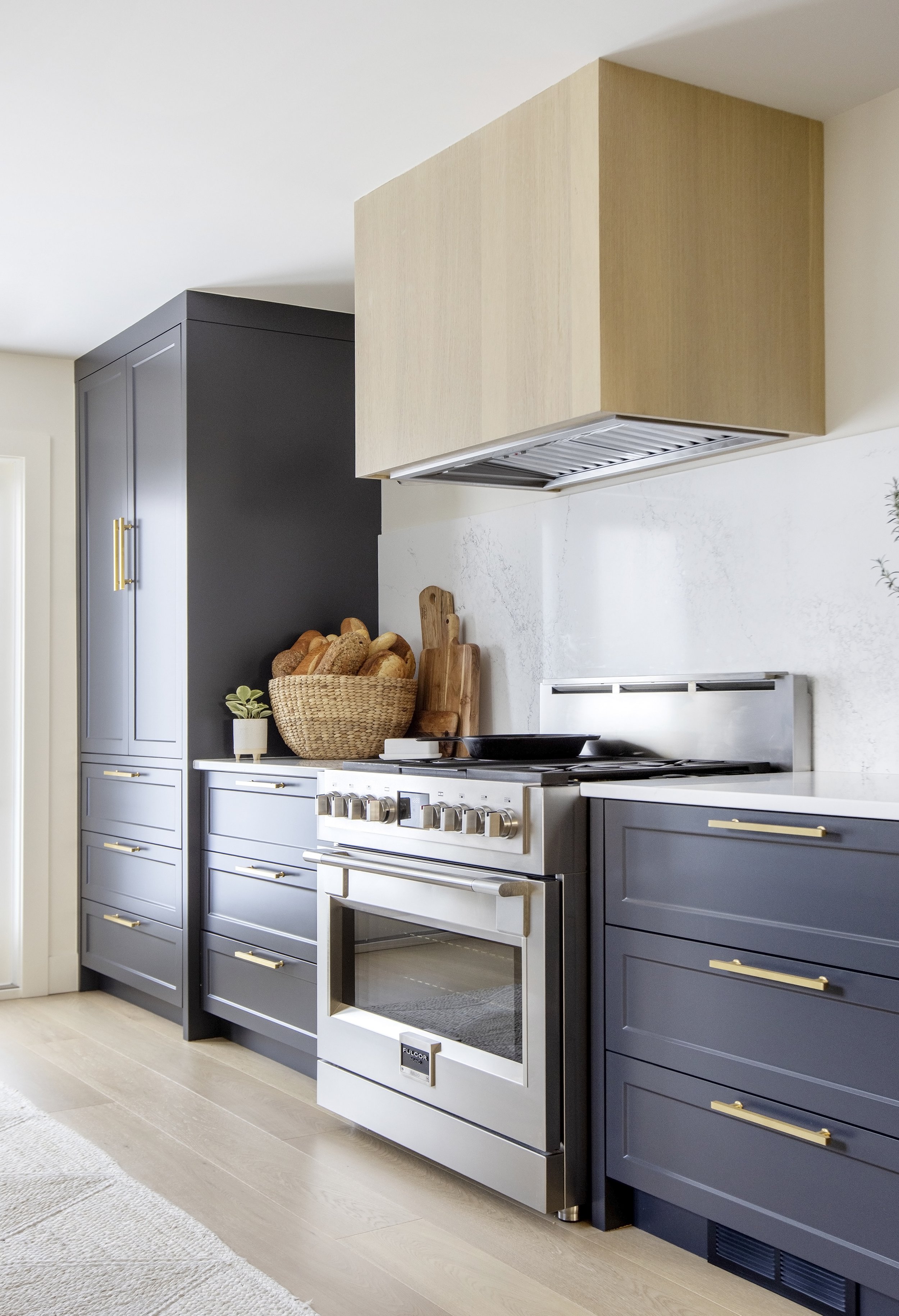 Modern kitchen with navy blue cabinets, a stainless steel oven and stove, a wooden range hood, and decor including bread basket and small potted plants.