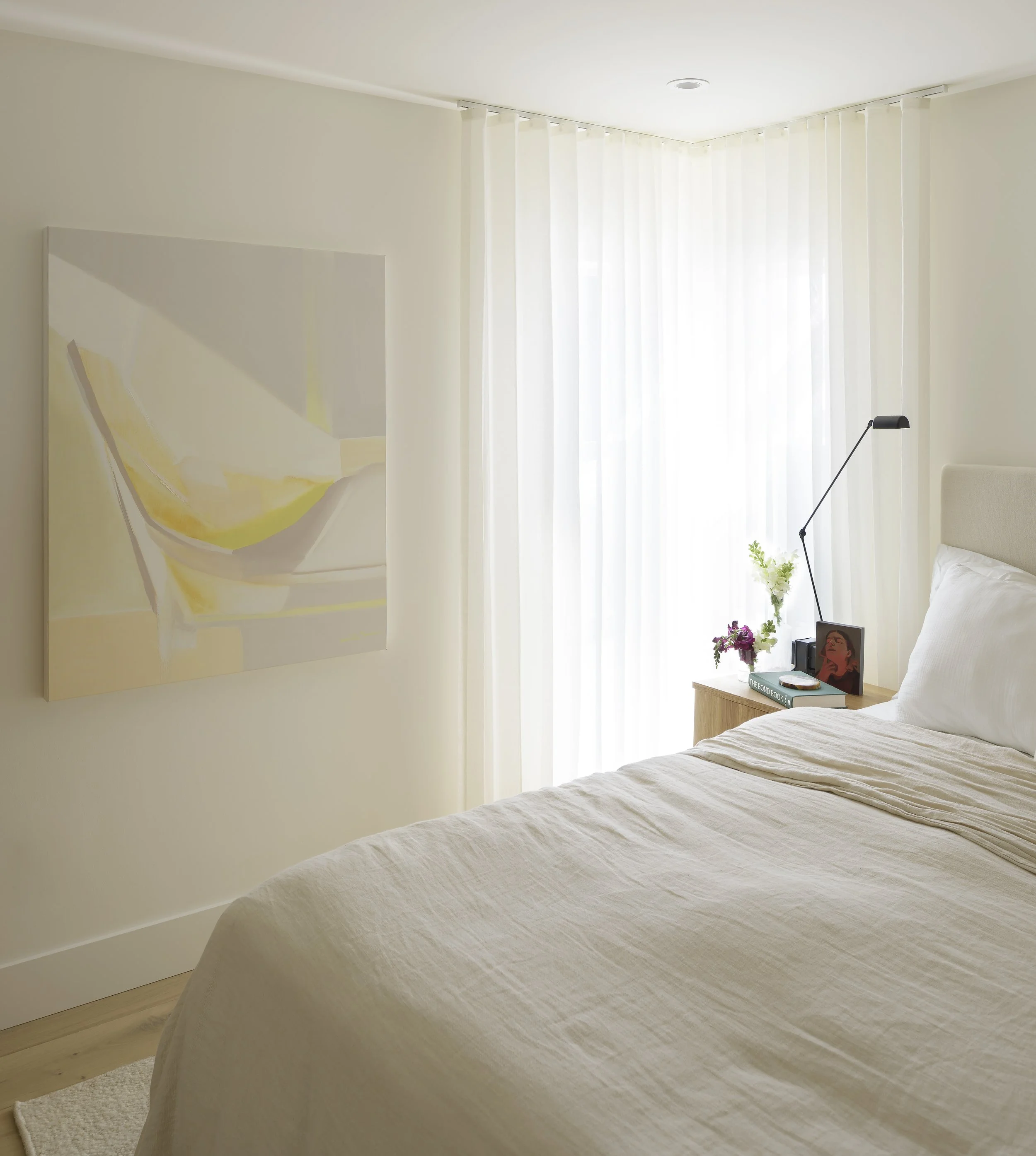A cozy bedroom with a neatly made bed covered in a light beige quilt, a nightstand with books and flowers, a framed portrait, a lamp, and abstract artwork on the wall. Large windows with sheer white curtains let in natural light.