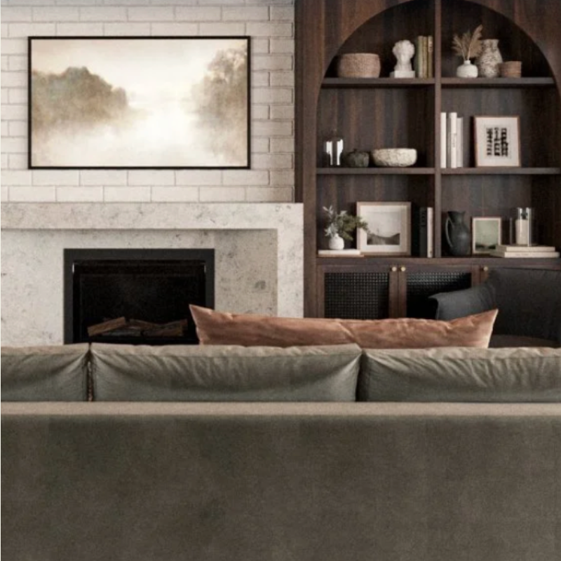 Living room with a gray sofa, a brown cushion, a fireplace with a white mantel, a framed landscape art piece above the fireplace, and a dark wooden bookshelf filled with decorative objects and books.