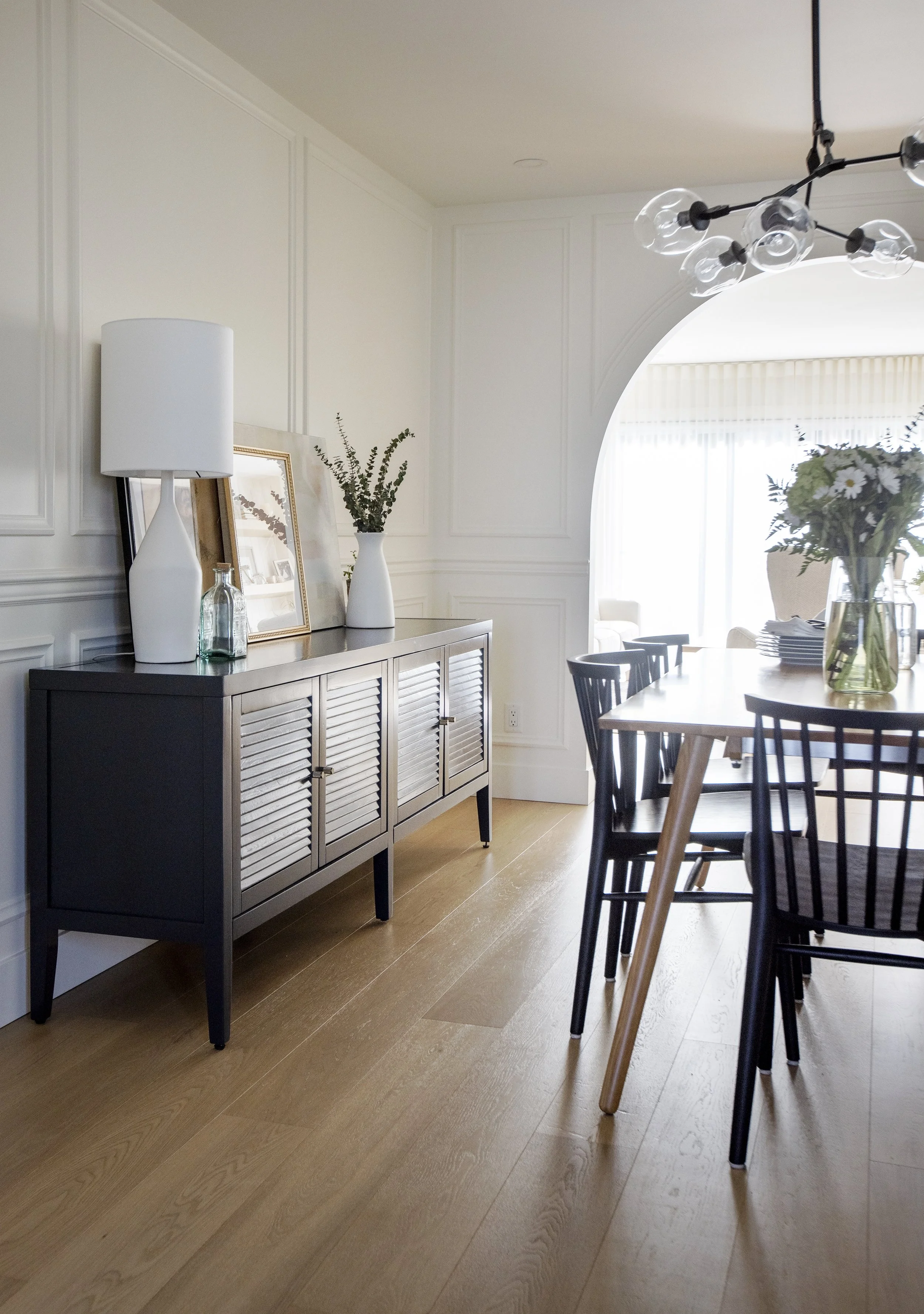 A corner of a modern dining room with a black and white sideboard, decorative vases with greenery, framed pictures, and a large chandelier hanging from the ceiling. A dining table with black chairs is partially visible.