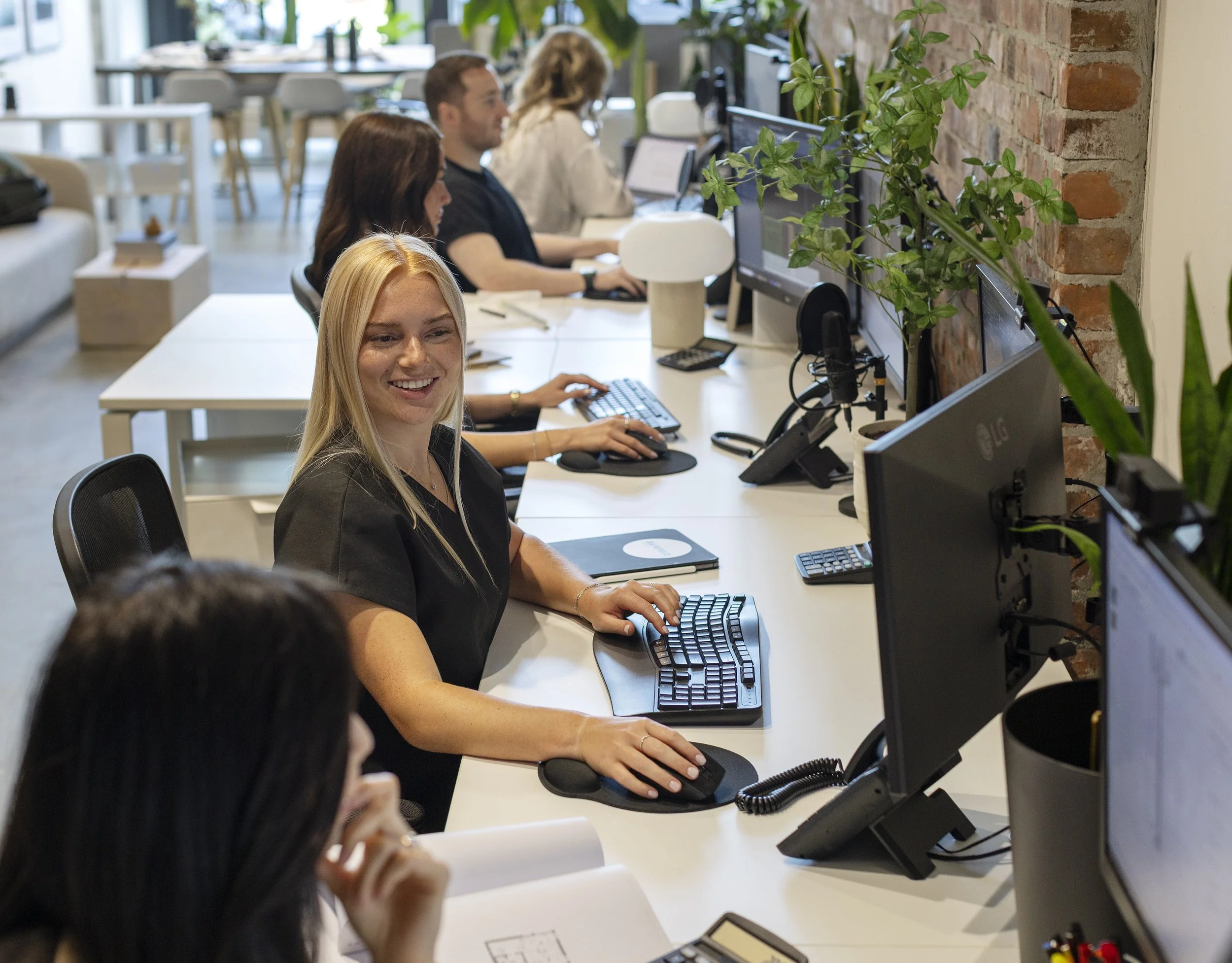 People working at desks with computers in a modern office with brick wall and green plants.