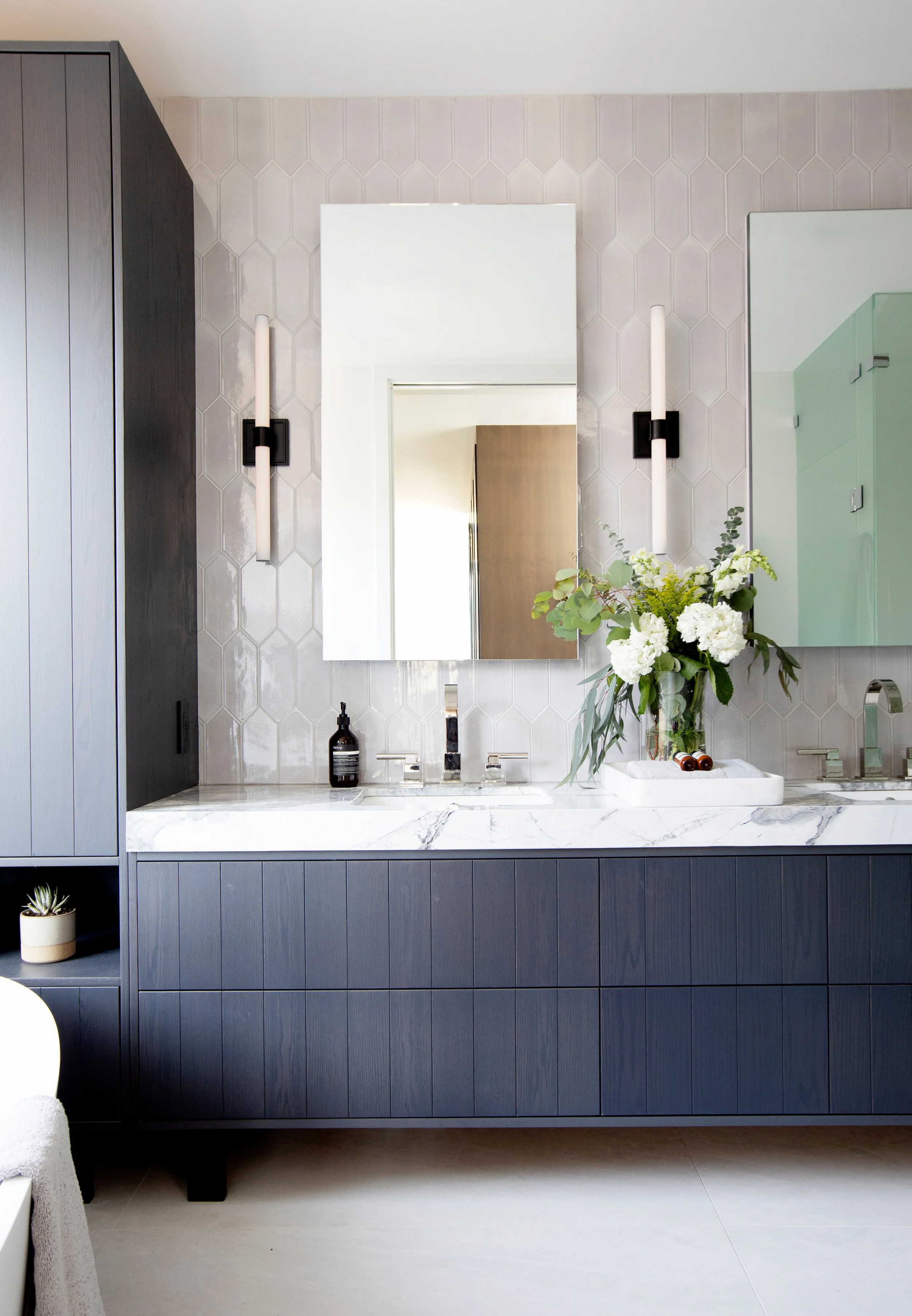 Modern bathroom with a blue cabinet vanity, marble countertop, wall mirror, wall-mounted light fixtures, a vase with white flowers, and various toiletries.