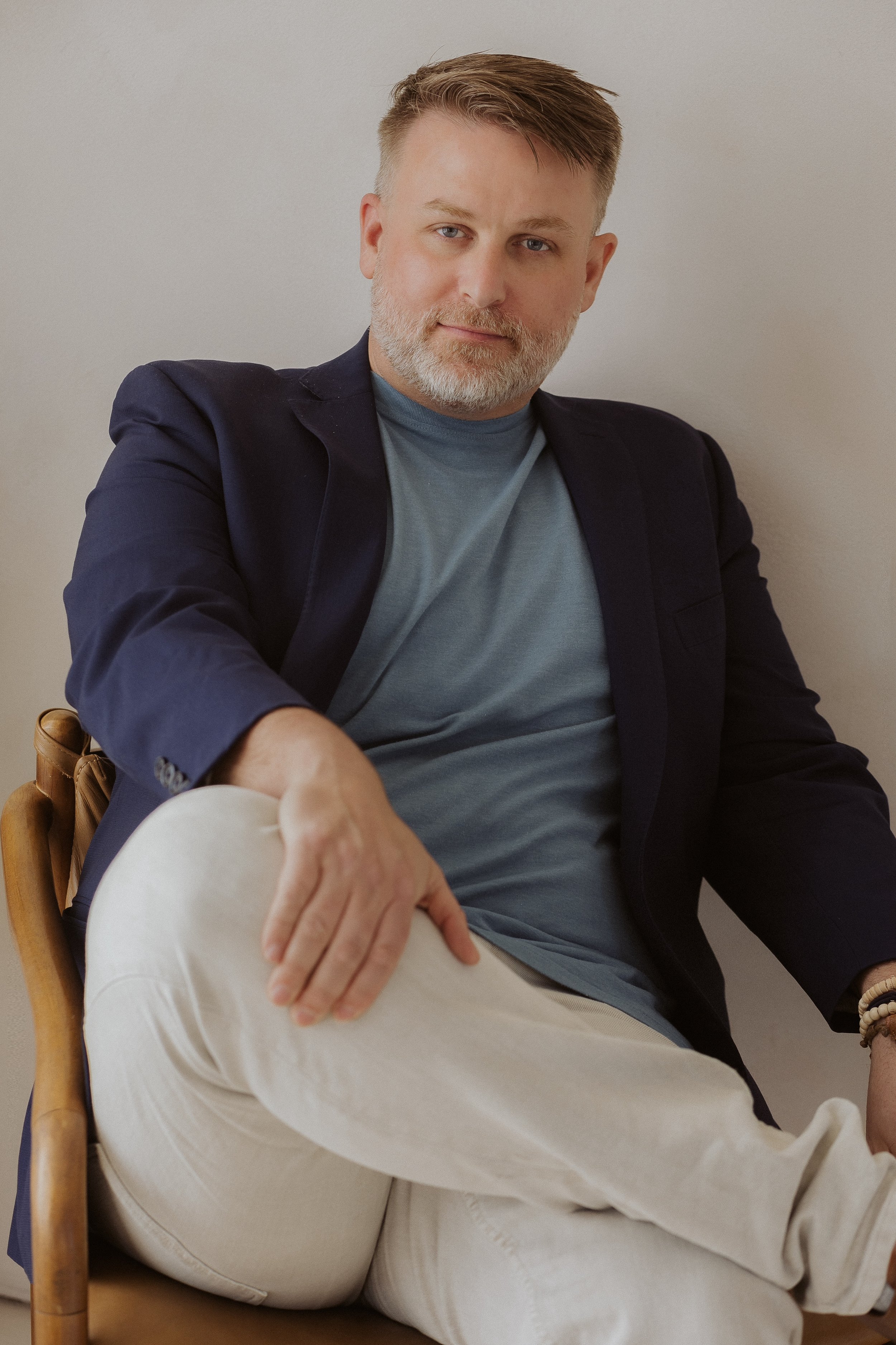 A man with gray beard and blue eyes wearing a dark blazer and blue shirt, sitting on a chair with a wooden frame, leaning back against a plain light-colored wall.
