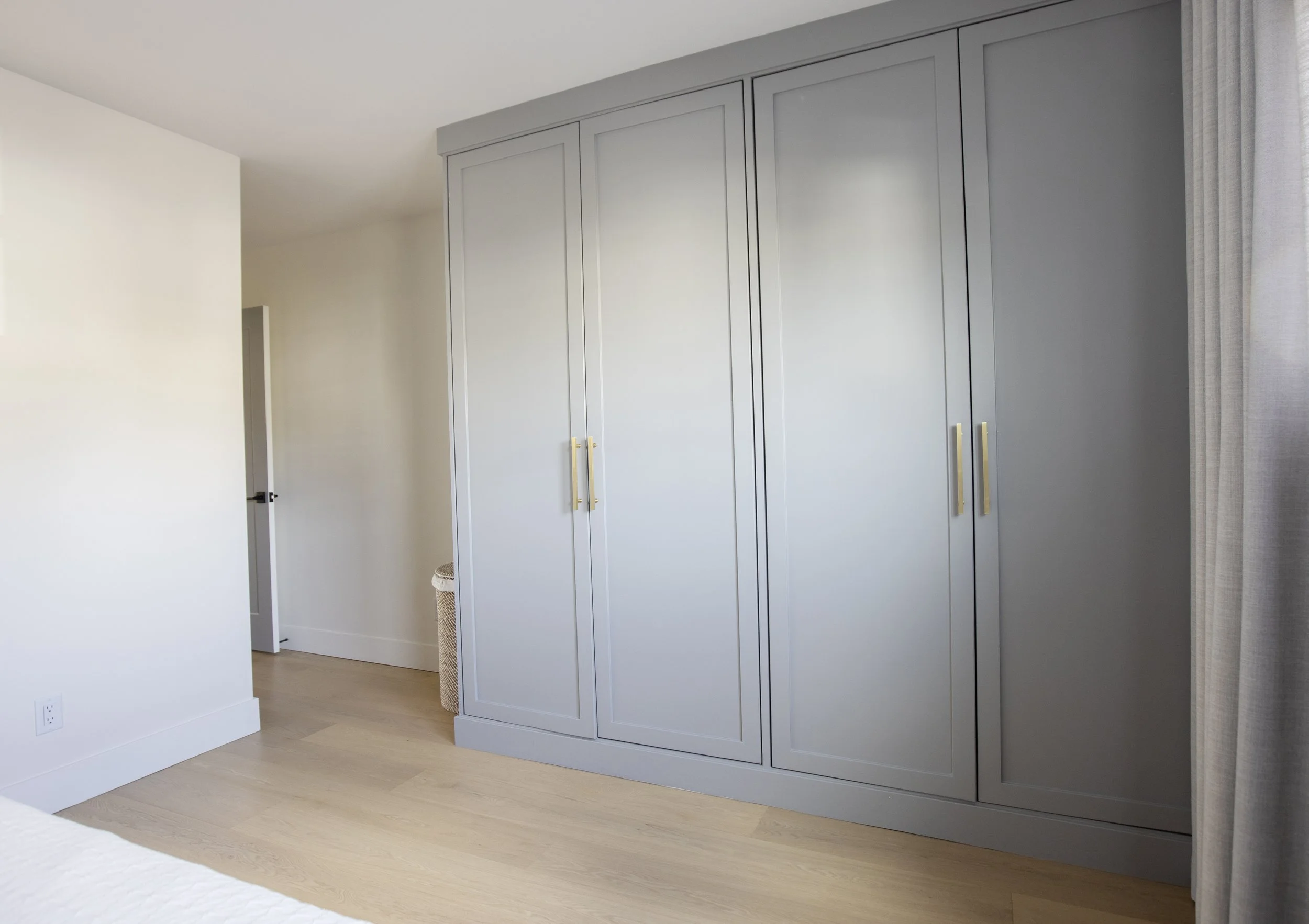 A bedroom with a white wall, a large gray closet with gold handles, and a light hardwood floor. There is a partially visible bed in the foreground with a white quilt. A woven laundry basket is positioned next to the closet. A white door is open on th