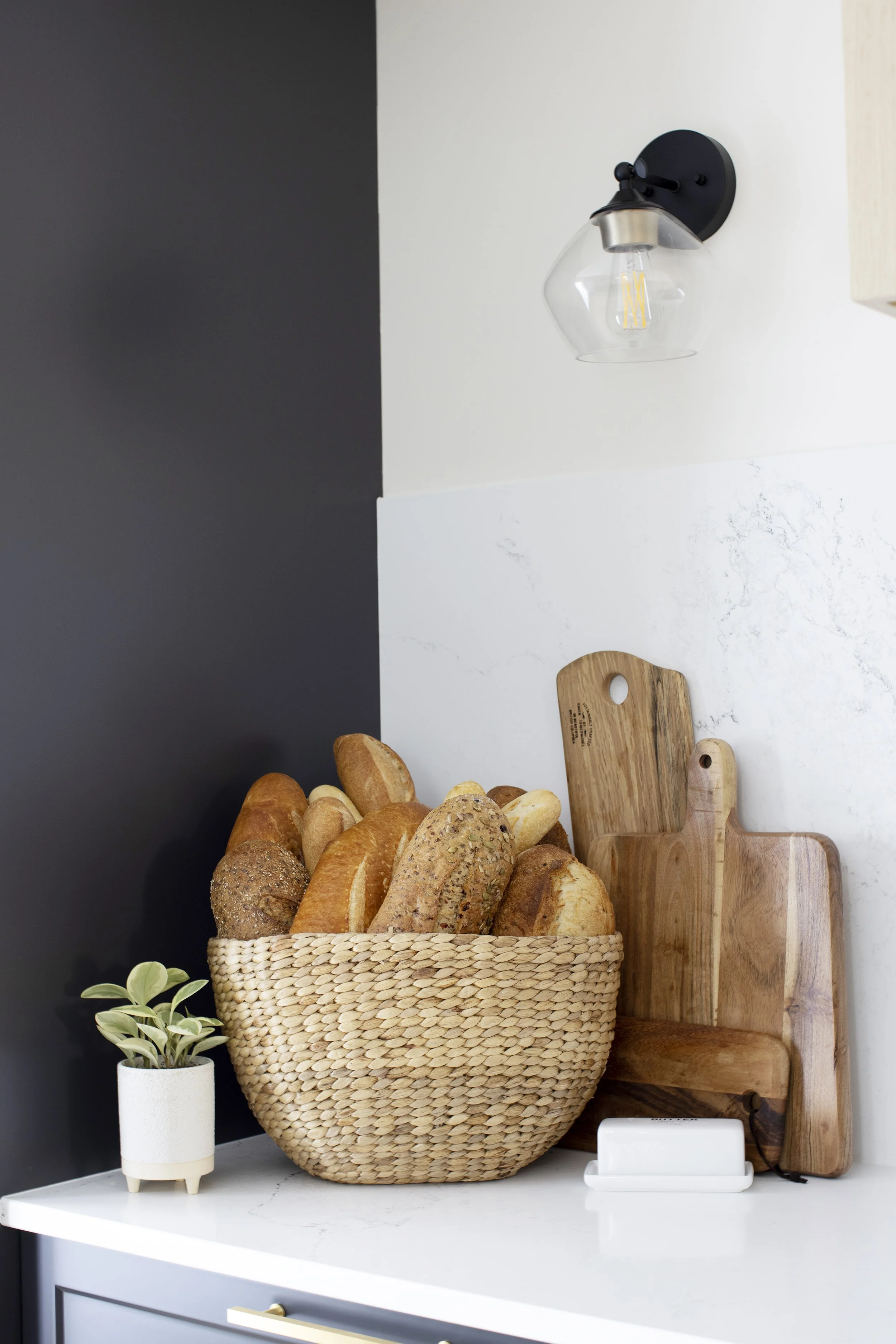 Basket filled with various types of bread on a white kitchen counter, with a small potted plant, wooden cutting boards, and a wall sconce light in the background.
