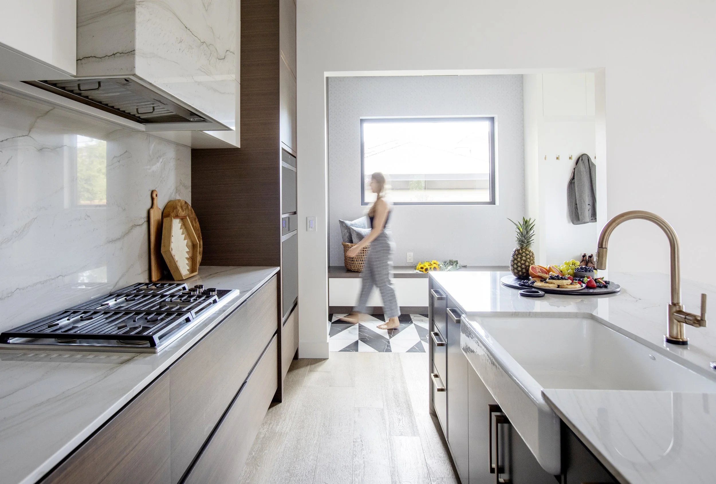 A modern kitchen with white marble countertops and backsplash, wooden cabinets, a woman walking past, and a window showing outdoor scenery. A fruit platter with pineapple, grapes, strawberries, and sliced fruit is on the counter.