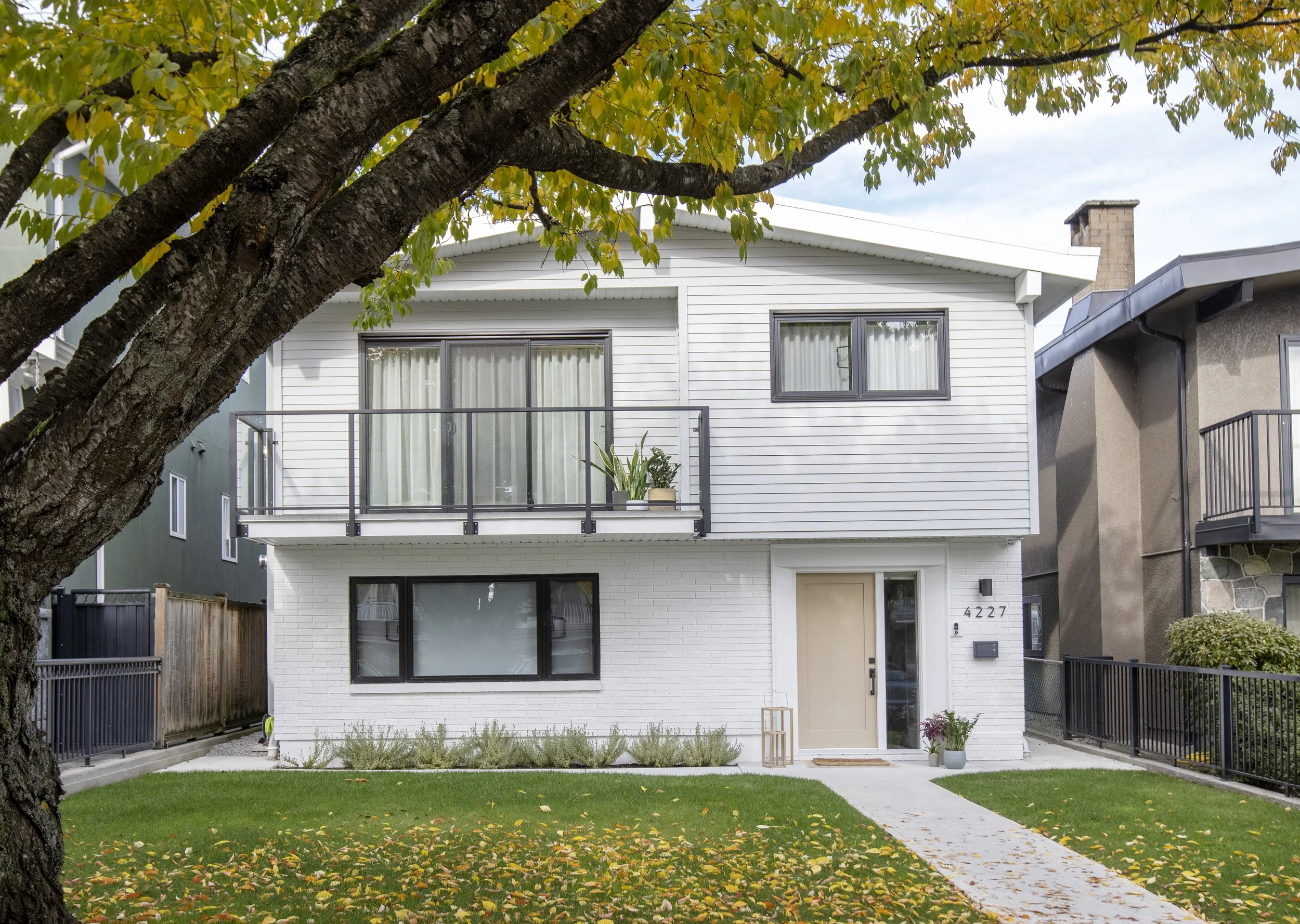 A modern white two-story house with a small front yard, a concrete pathway, and autumn leaves scattered on the grass. The house has large windows, a balcony with a potted plant, and is partially obscured by a large tree with green leaves.