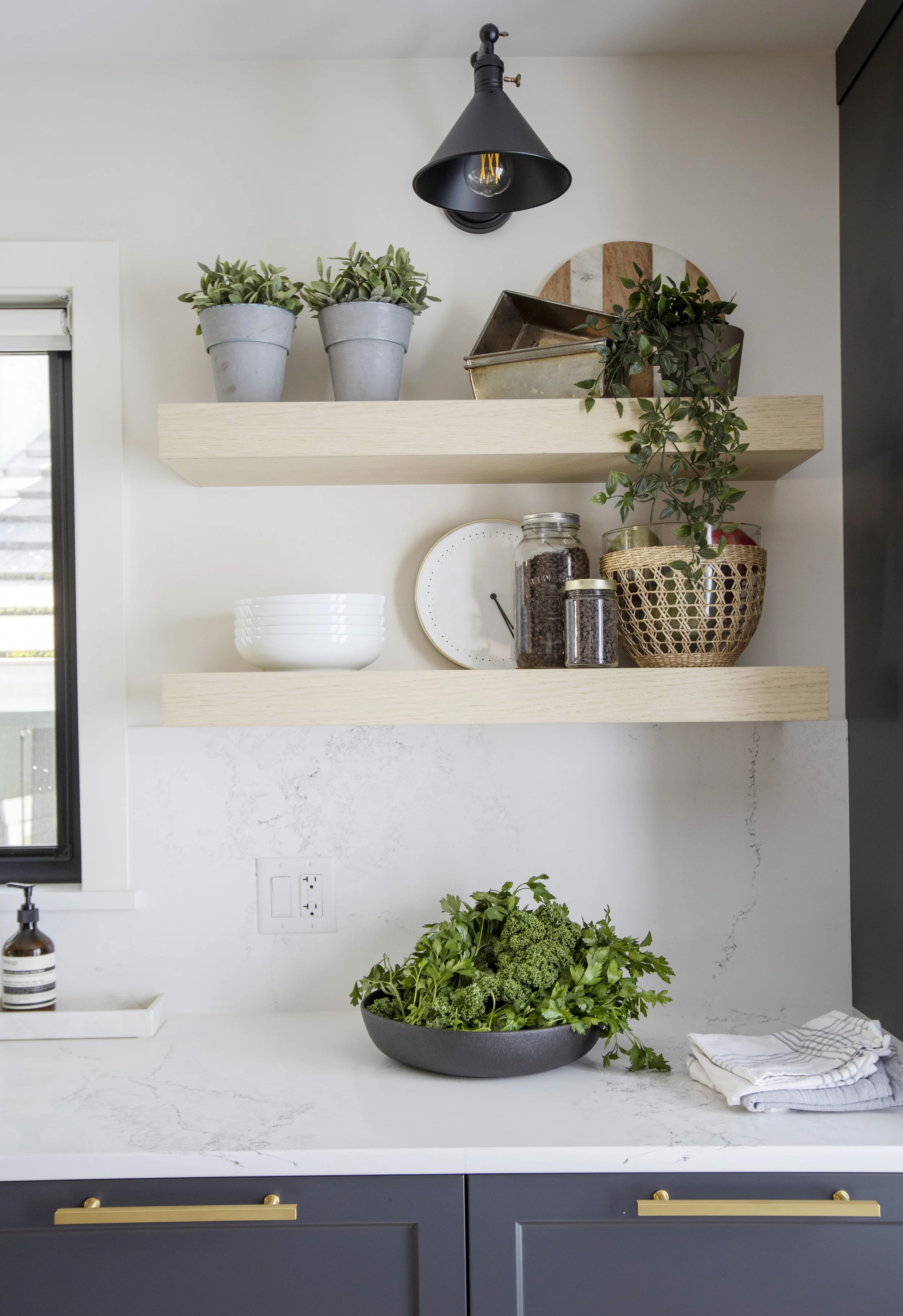 A modern kitchen with a white marble countertop, navy blue cabinets with gold handles, and open wooden shelves displaying potted plants, jars of coffee beans, and a decorative tray. A large bowl of fresh greens and a folded dish towel are on the coun