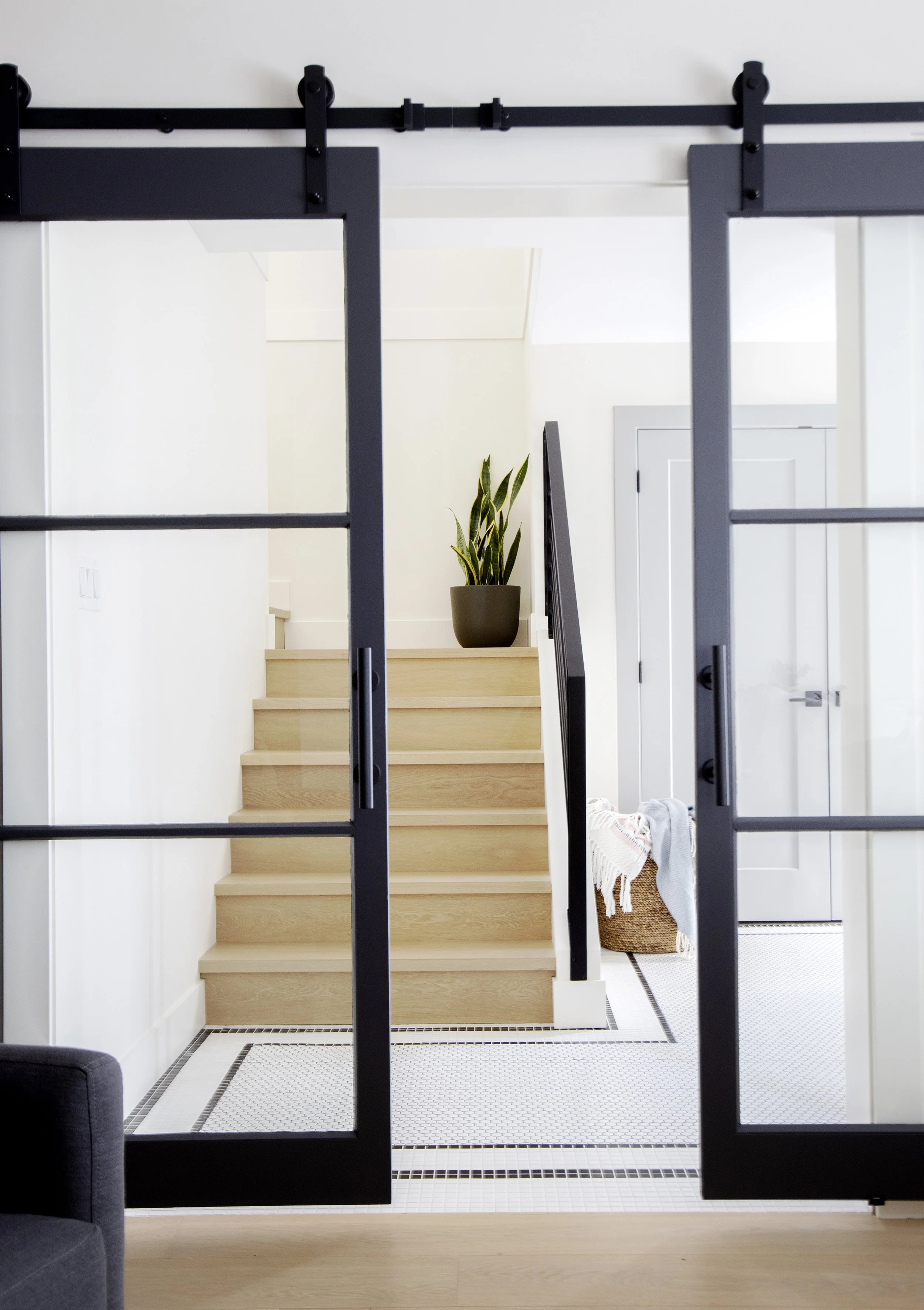 Interior view of a modern home with a black sliding barn door on a rail system, light wood stairs leading to an upper level, a potted plant on the landing, and a small basket with a blanket near a white door.