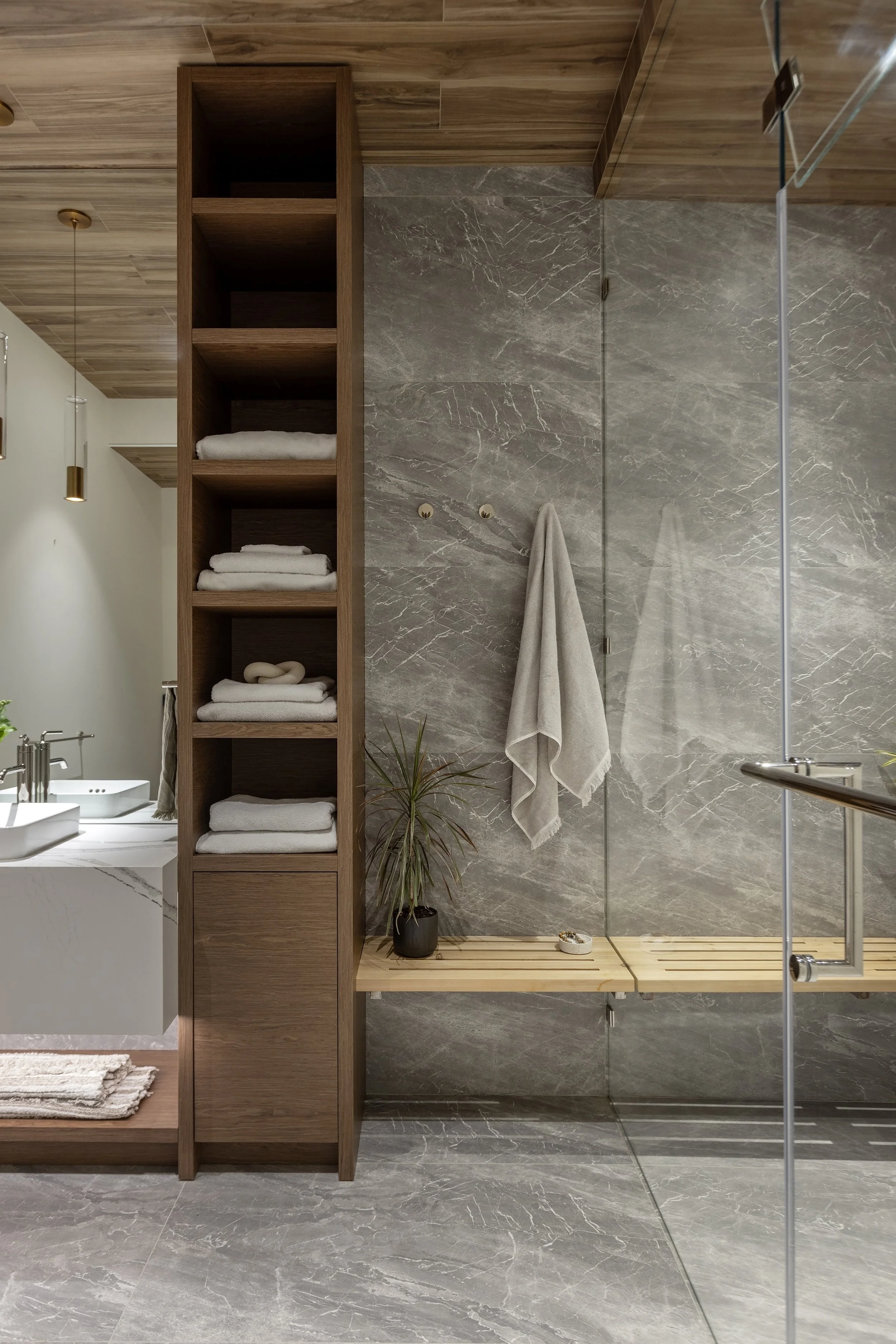 Modern bathroom with wooden shelving unit, gray marble wall, glass shower door, towel hanging on wall, potted plant, and wooden slatted shower bench.