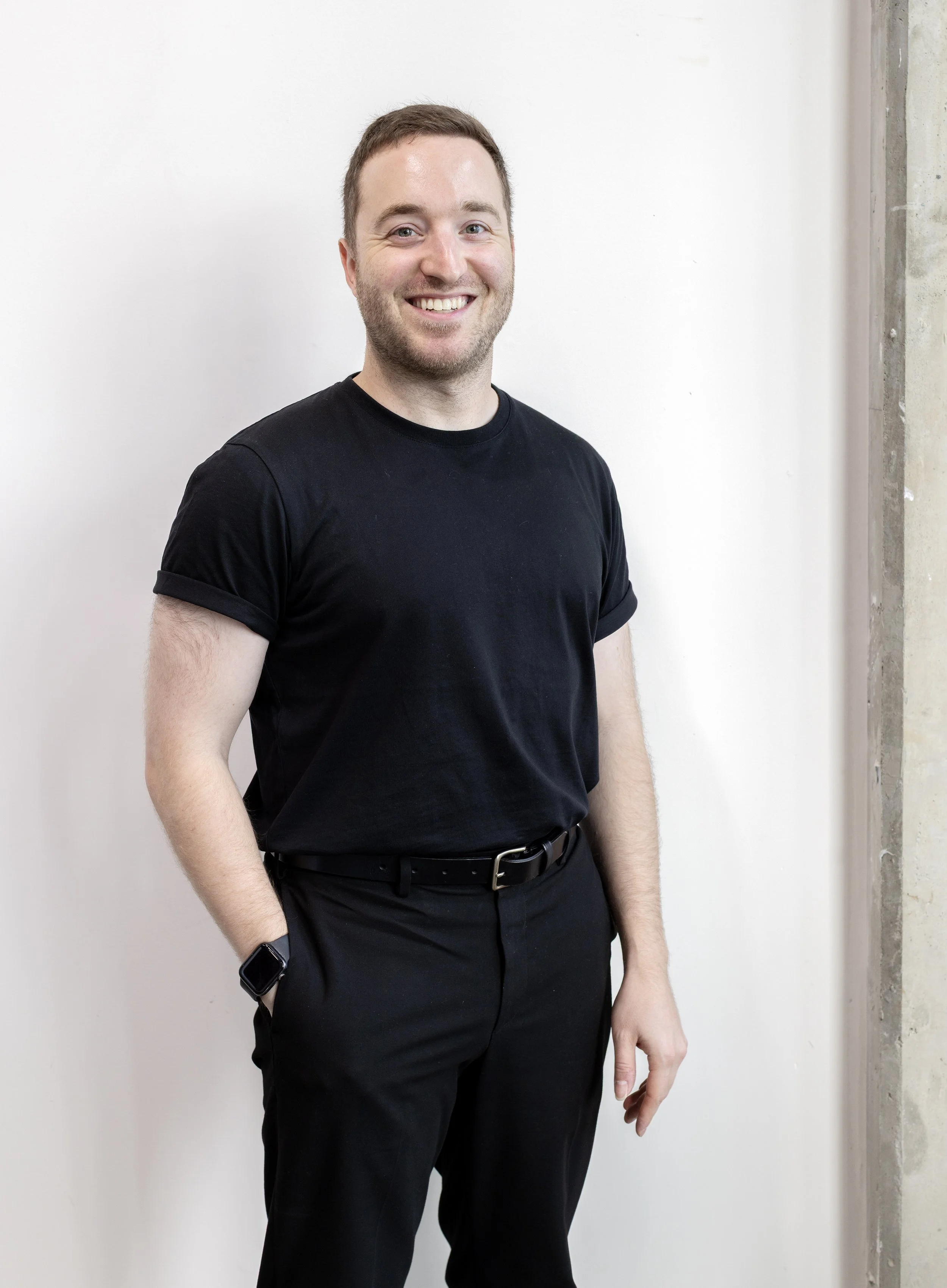 A young man with short brown hair and a beard, smiling, wearing a black T-shirt, black pants, a black belt, and a smartwatch, standing against a white wall with a concrete edge on the right.