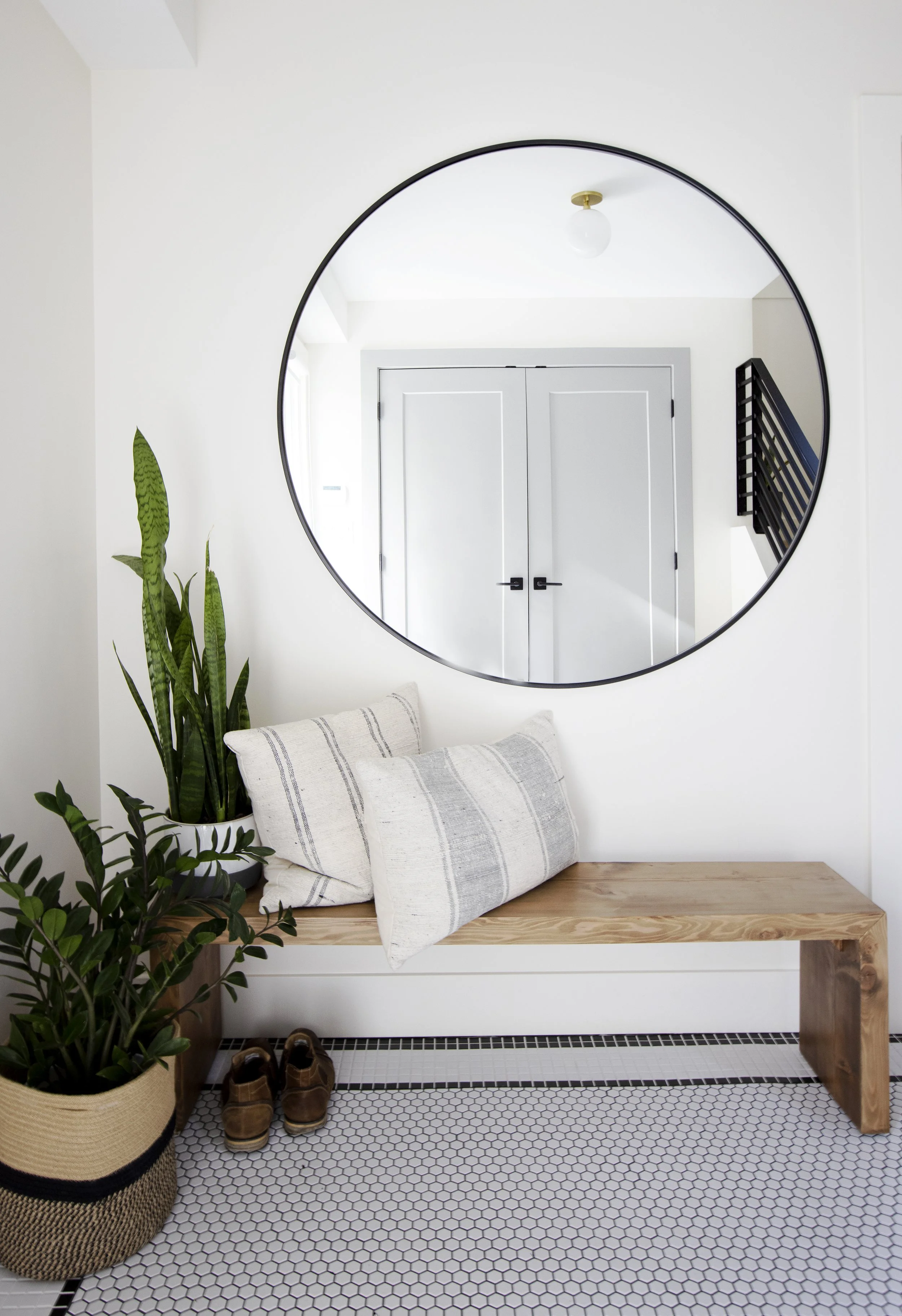 Entryway with a large round mirror on a white wall, a wooden bench with a striped pillow, a potted plant, and shoes on a hexagonal tile floor.