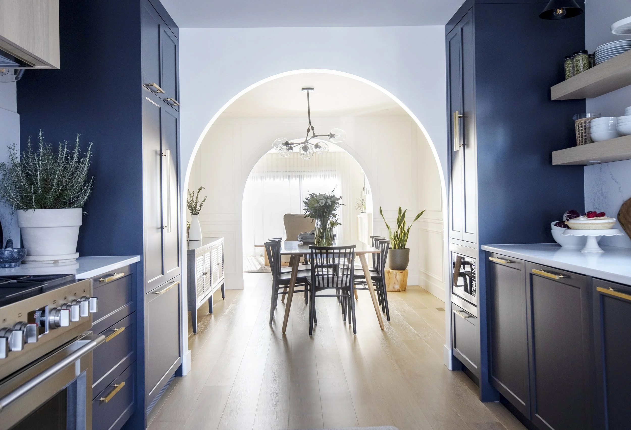 A view through a kitchen doorway showing a dining area with a table and chairs, decorated with vases and flowers. The kitchen has blue cabinets, open shelves with dishes, and a variety of plants.