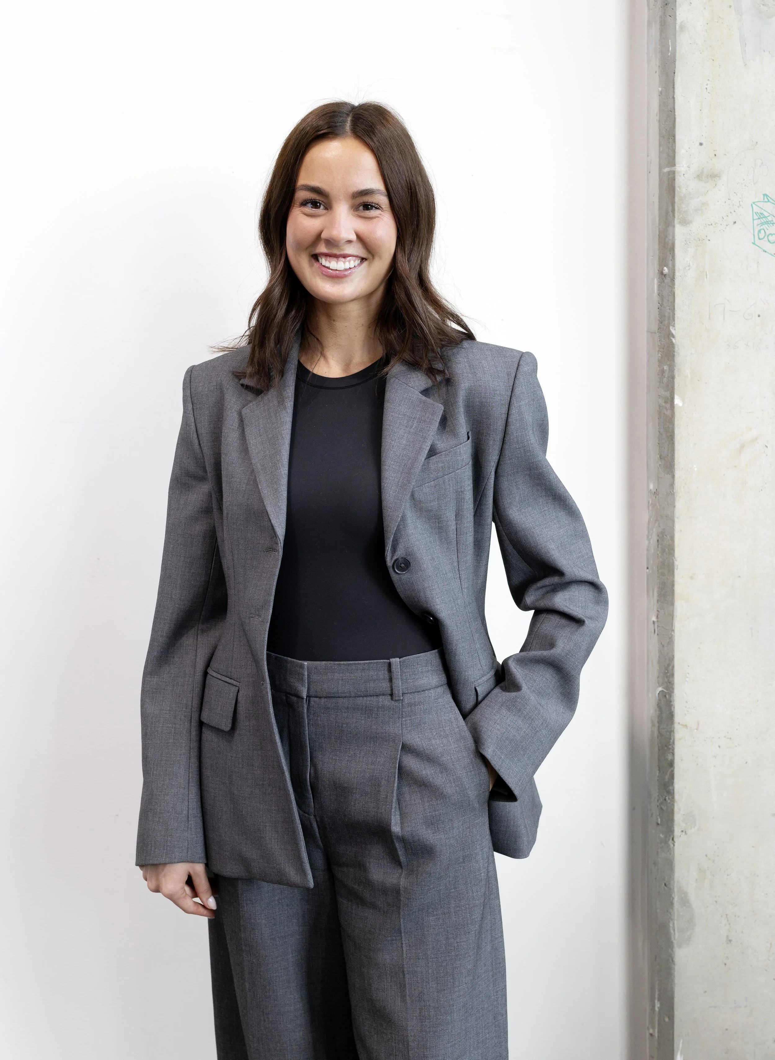 A young woman with shoulder-length brown hair, smiling, dressed in a gray business suit and black top, standing against a white wall with parts of a concrete wall visible on the right.