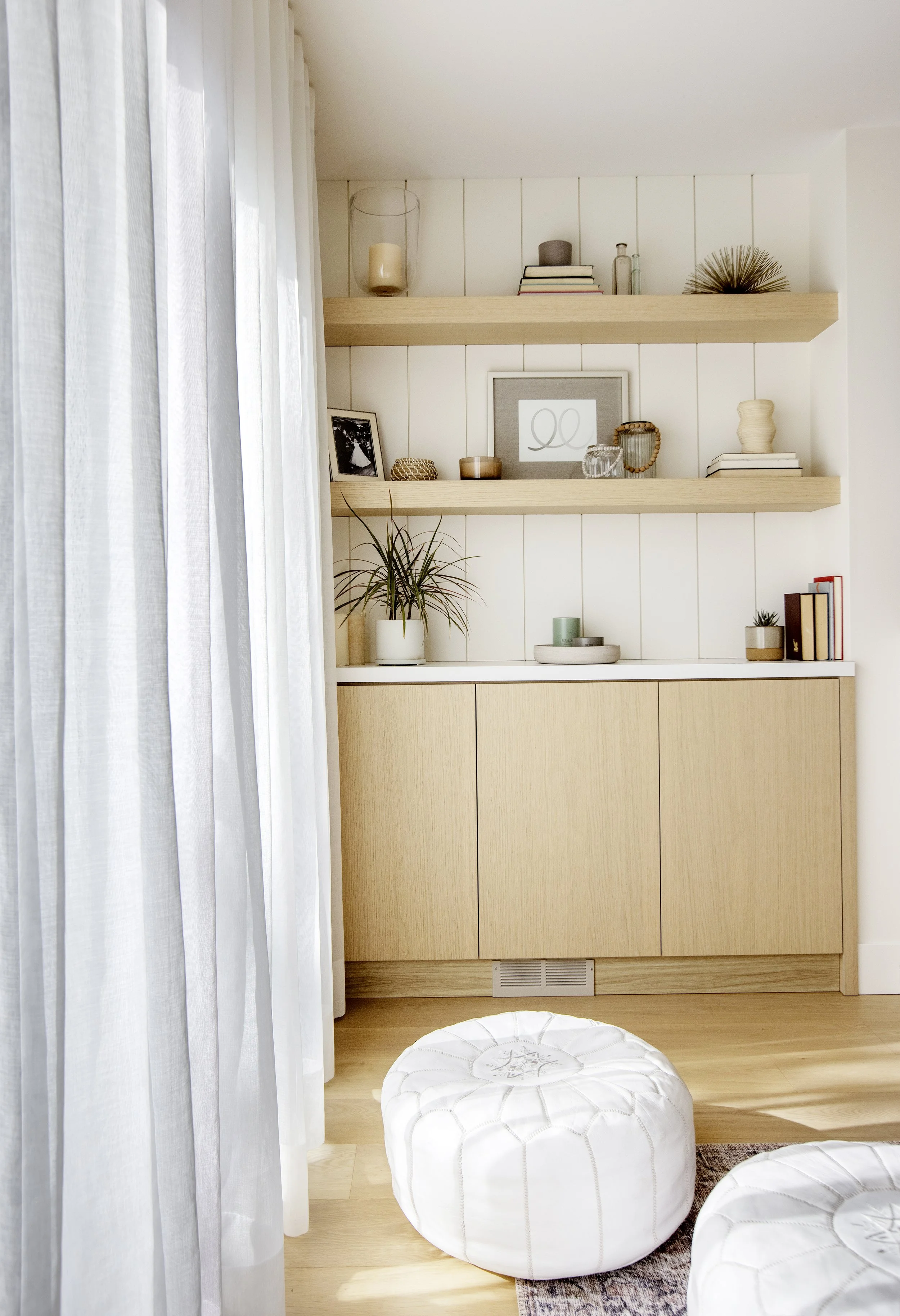Interior scene with beige storage cabinet, wooden shelves decorated with books, candles, framed photo, small plants, decorative objects, and sheer white curtains on the left side of the image.