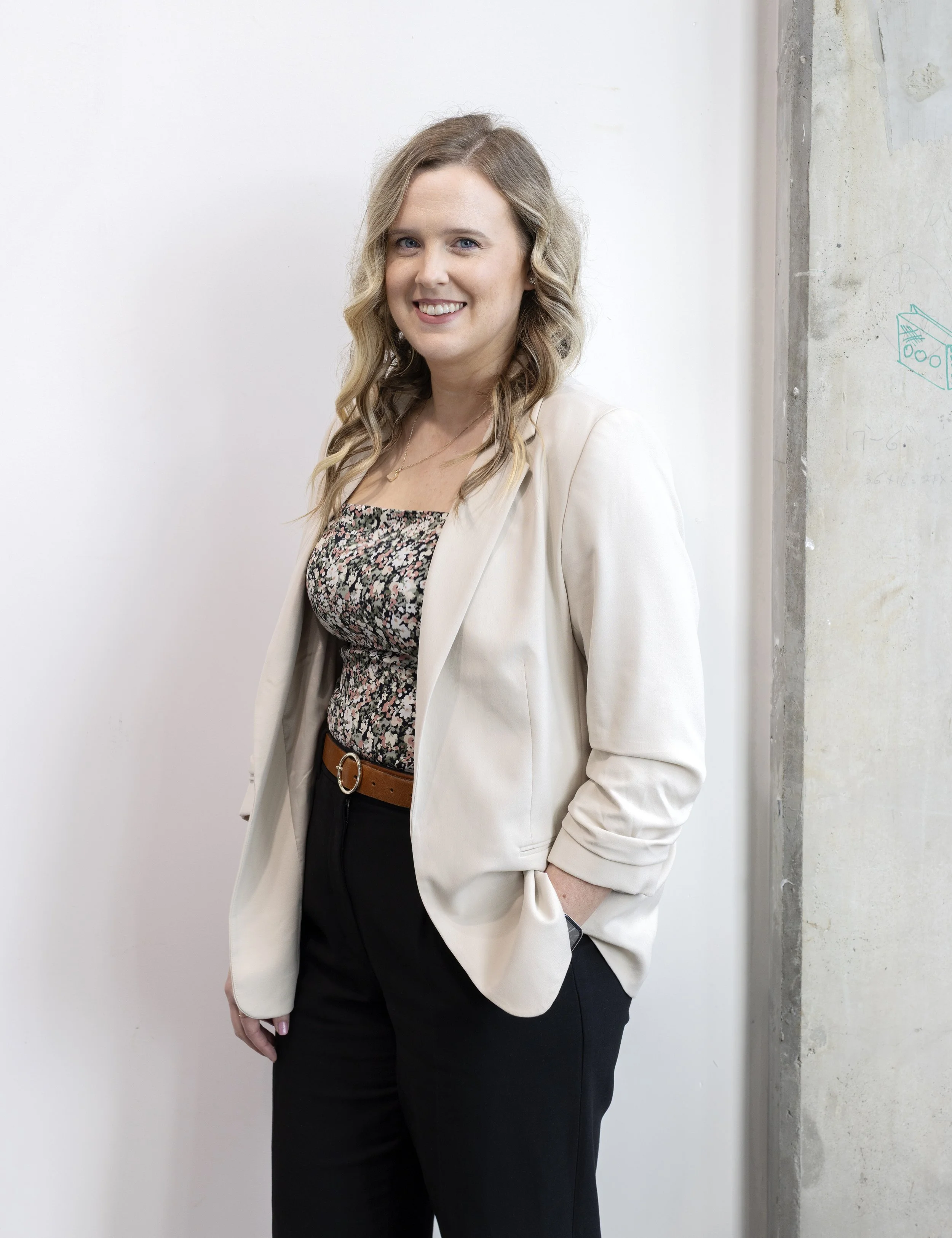 A woman standing against a white wall and a concrete pillar, wearing a cream blazer, floral blouse, and black pants.