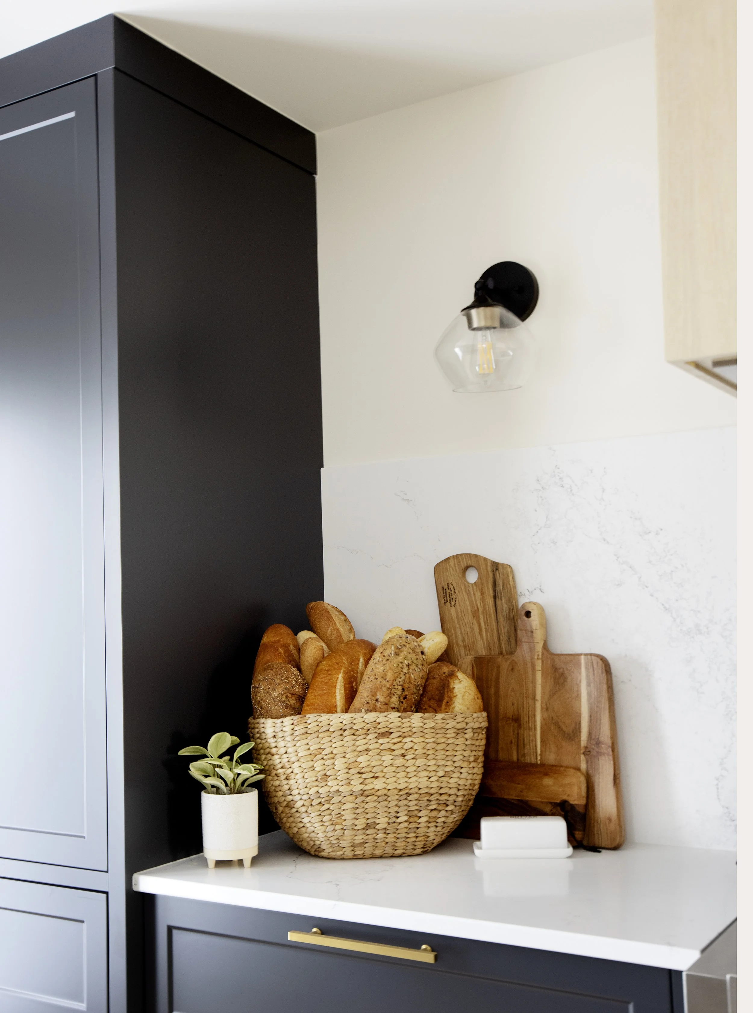 Kitchen countertop with a woven basket filled with various breads, a small potted plant, and wooden cutting boards against a white wall and black cabinetry with a gold handle.