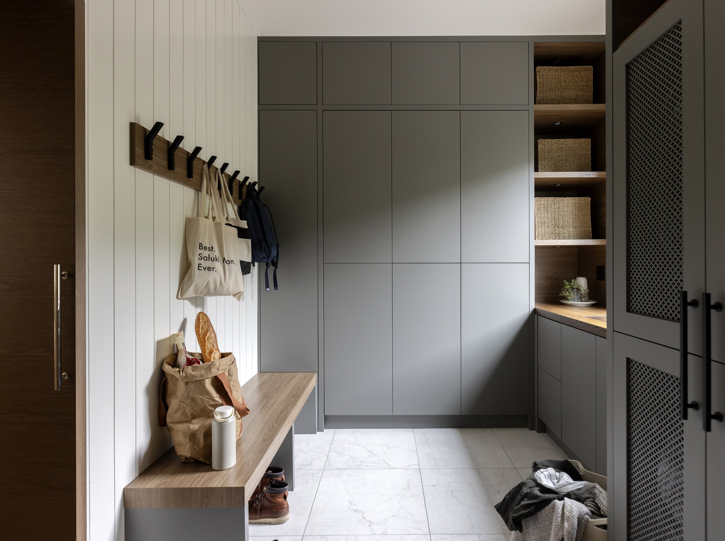 A modern entryway with gray cabinets, a wooden bench, and wall hooks holding bags and clothing. There are storage baskets on open shelves, shoes on the floor, and a small plant on a shelf. The floor is white tile, and a door is partially visible on the left.