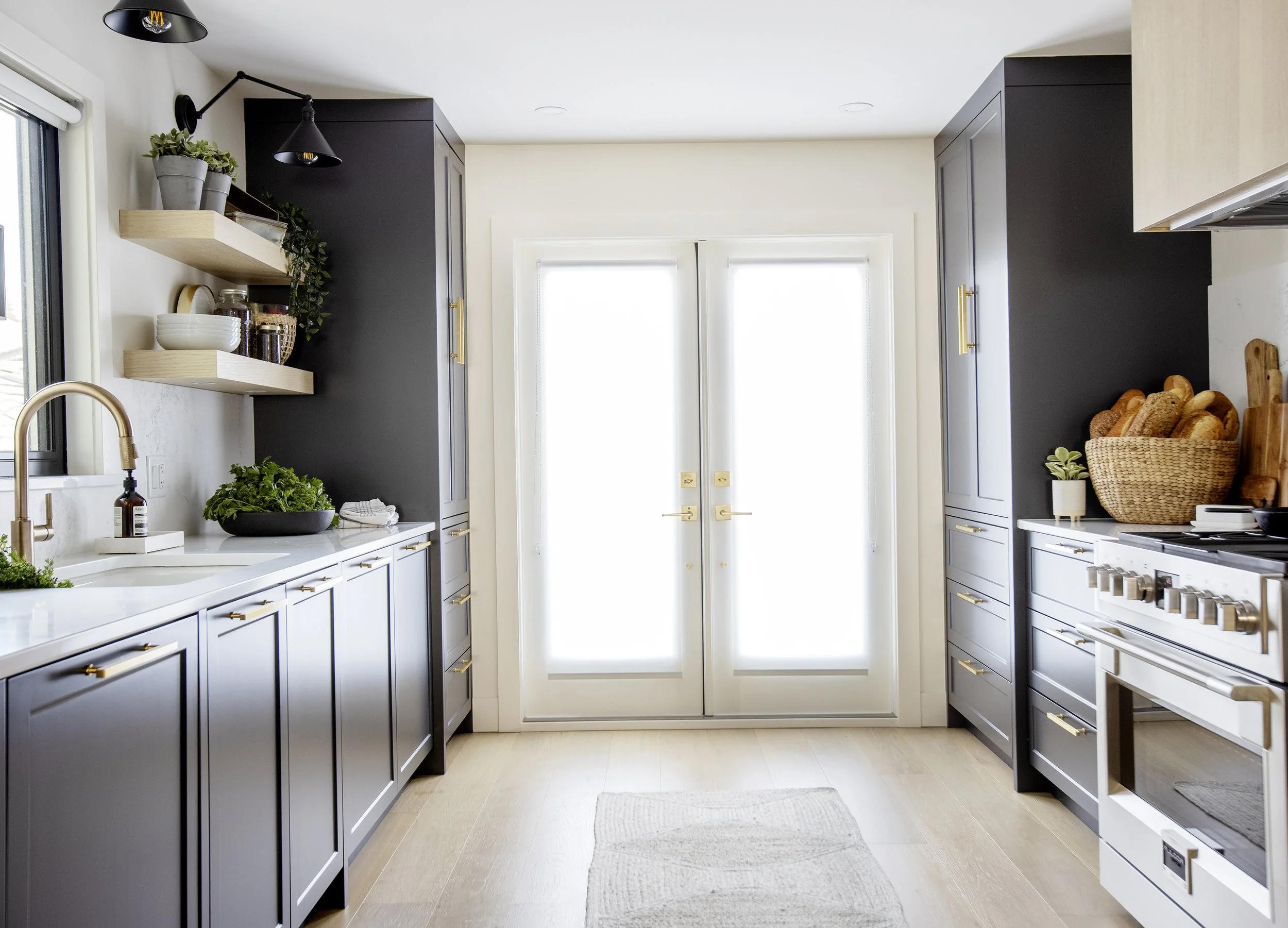 A modern kitchen with dark cabinets, white countertops, and open shelves with plants and dishes. Double glass doors let in natural light, and a rope basket with bread is on the right.