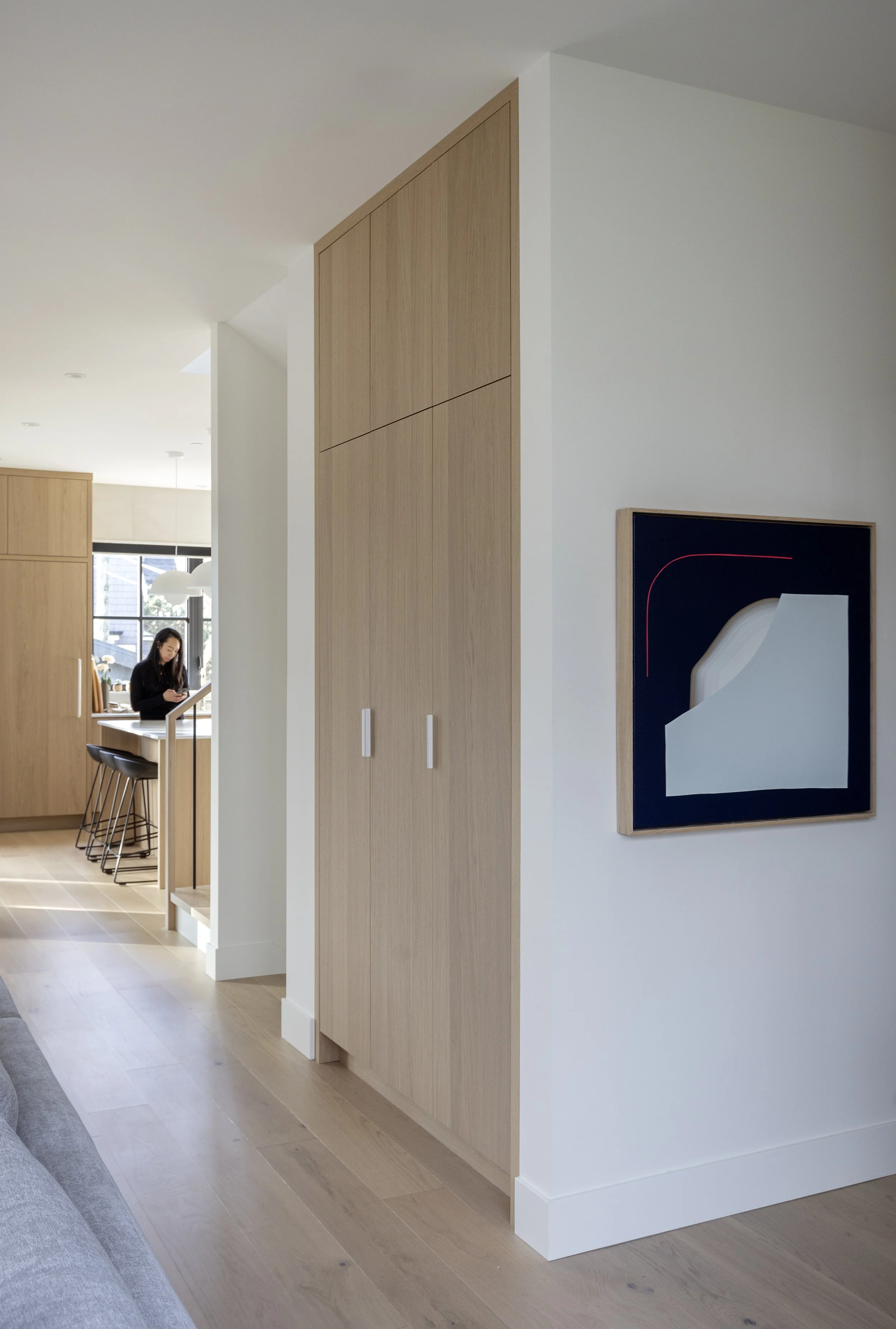 Living space with wooden cabinetry and modern art on the wall, a woman in dark clothing standing by a kitchen island with bar stools, natural light coming through windows, and light-colored wooden floors.