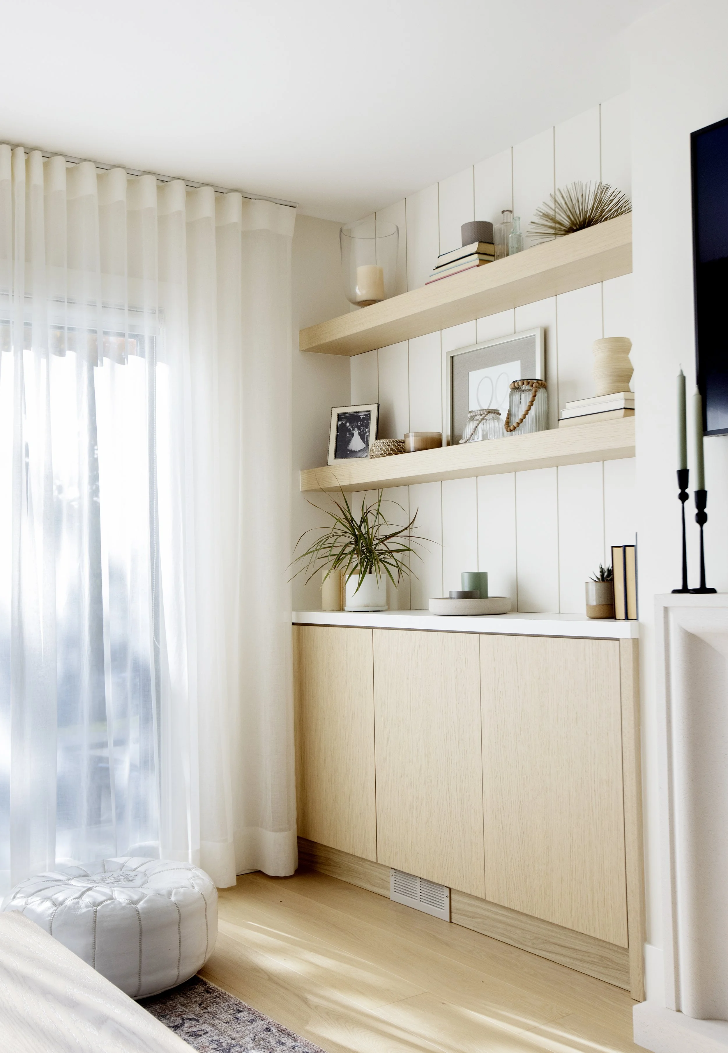 Bright living room with light wood cabinets, floating shelves with decor, a large window with sheer white curtains, and a white tufted ottoman on a patterned rug.