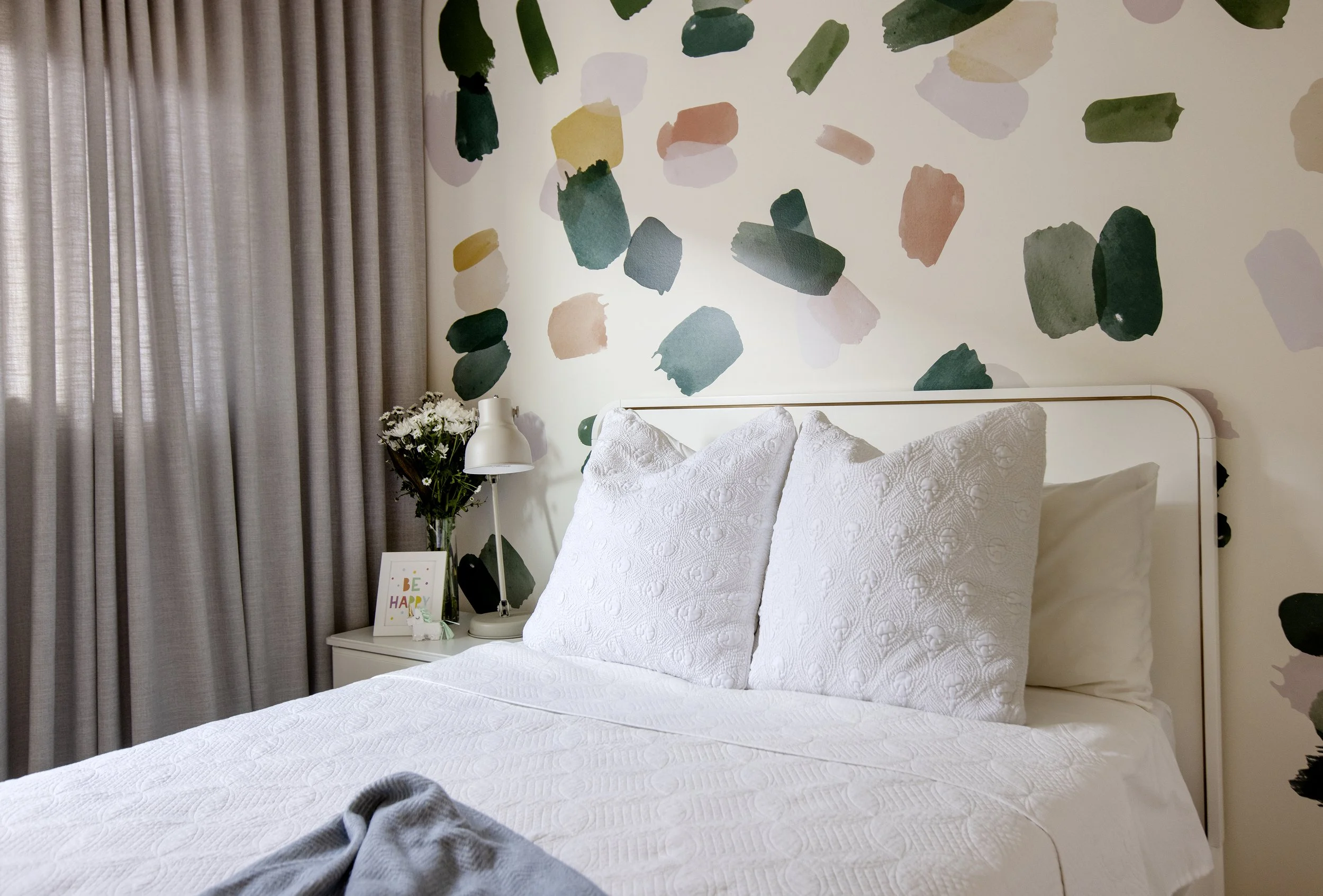 A bedroom with a white bed, white textured bedding, and pillows, next to a white nightstand with a vase of white flowers, a white lamp, and a small framed picture. The wall behind the bed has abstract watercolor wall art in shades of green, yellow, a