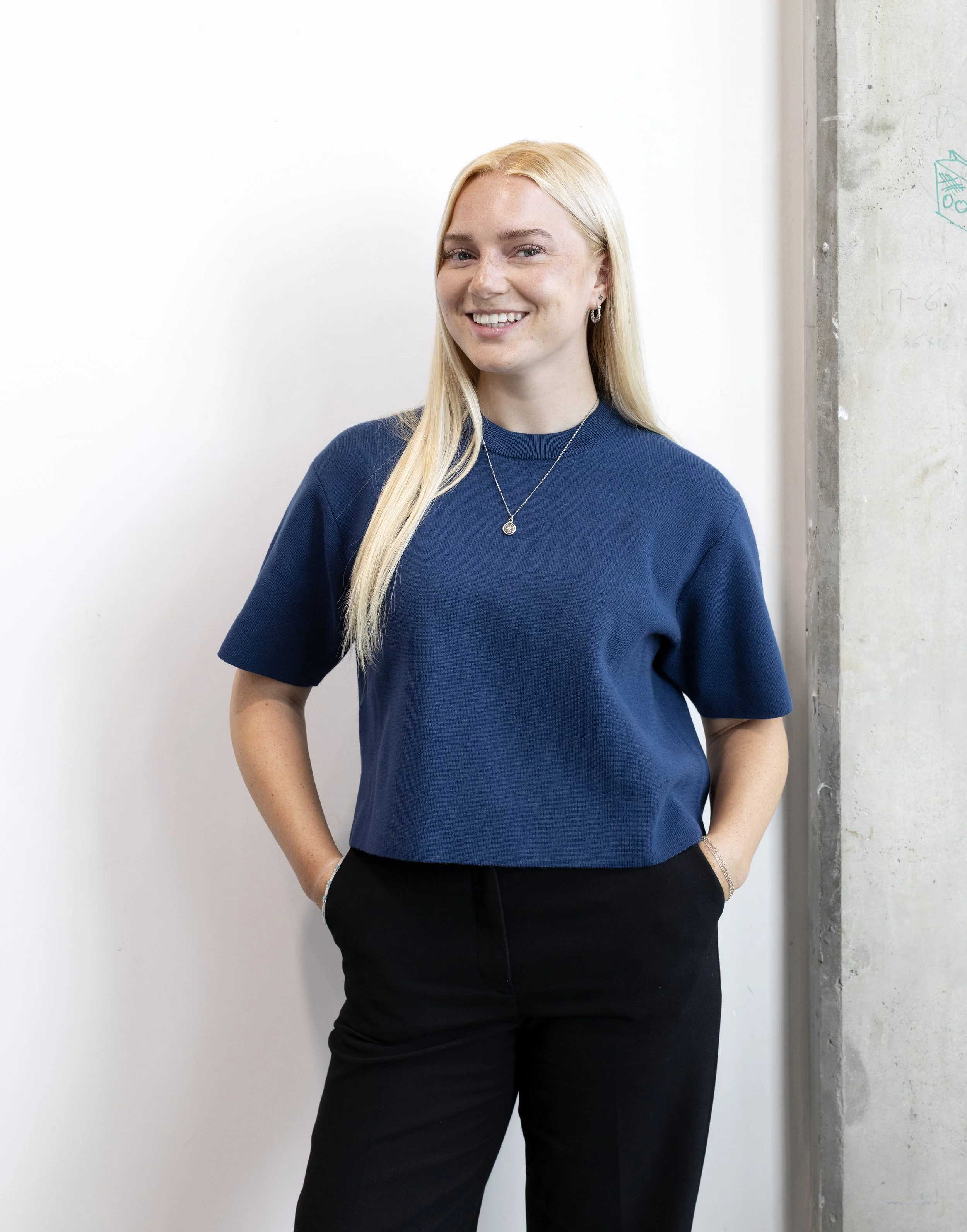 A woman with long blonde hair, wearing a blue top and black pants, standing indoors against a white wall and a concrete wall.