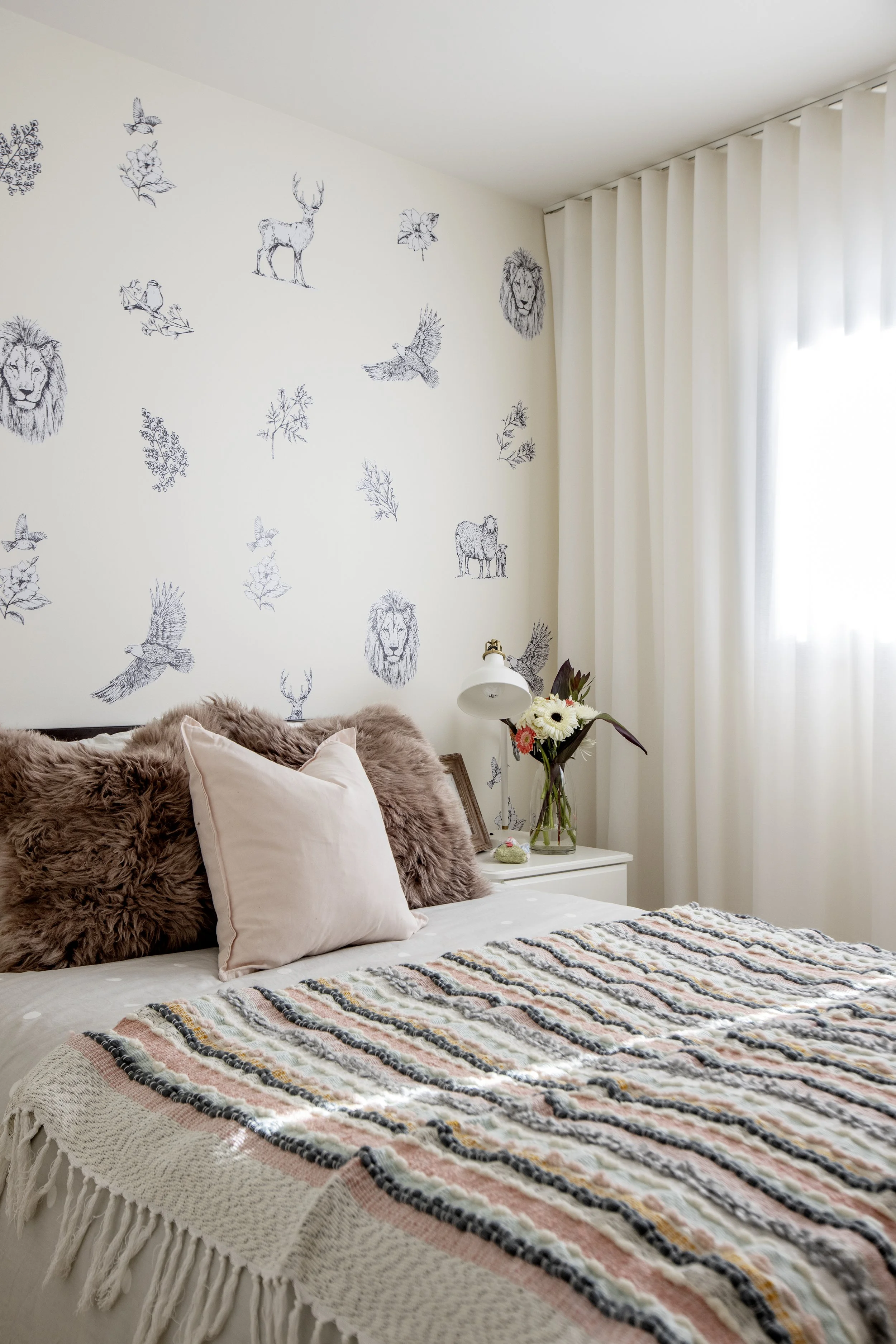 A cozy bedroom with a bed decorated with beige, pink, and black striped blanket, and fluffy pillows in brown and light pink. The wall behind has black line art animal and nature illustrations including lions, deer, birds, and leaves. A bedside table 