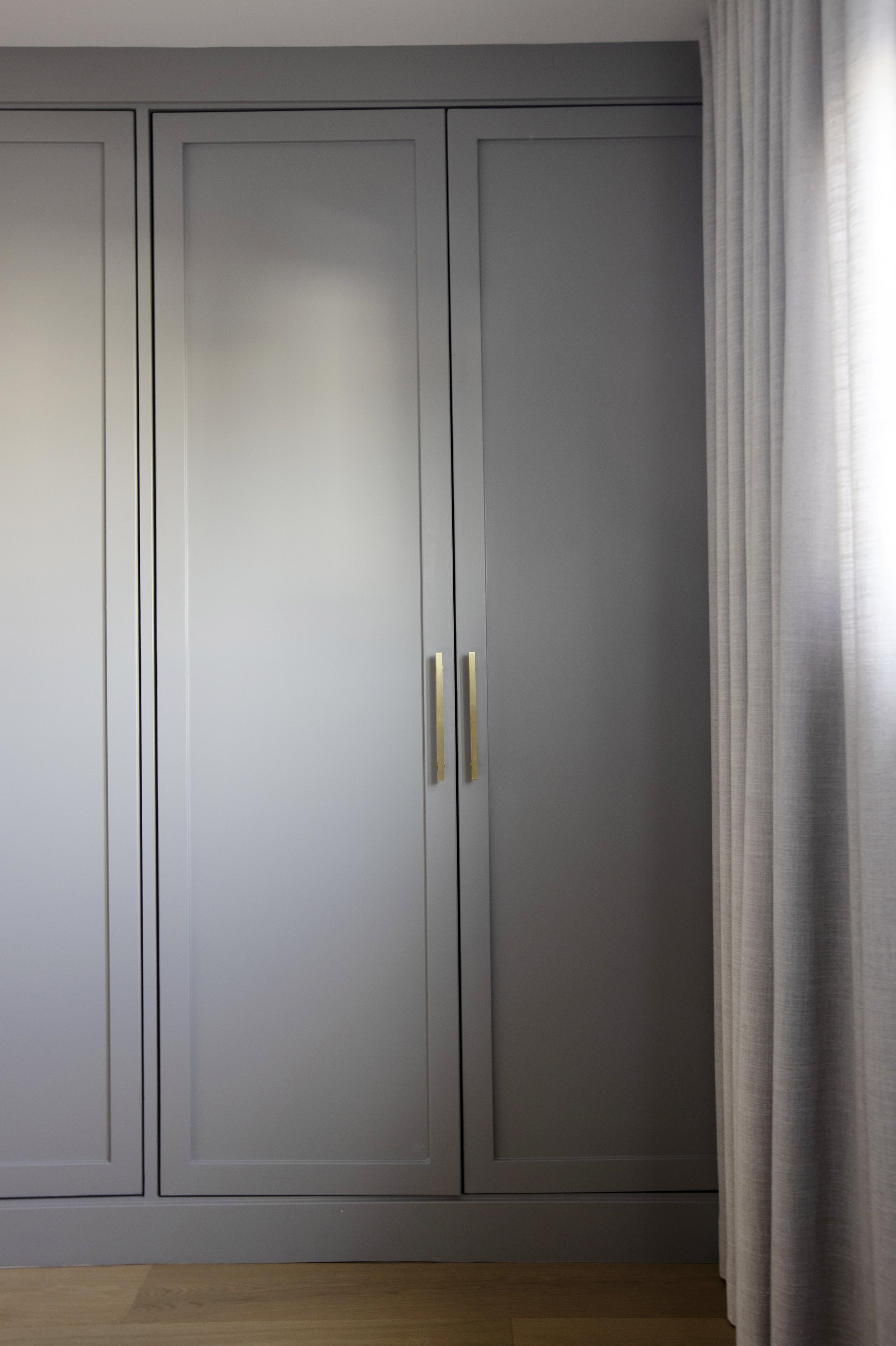 A gray wardrobe with three closed doors and gold handles, located next to light-colored curtains in a room.