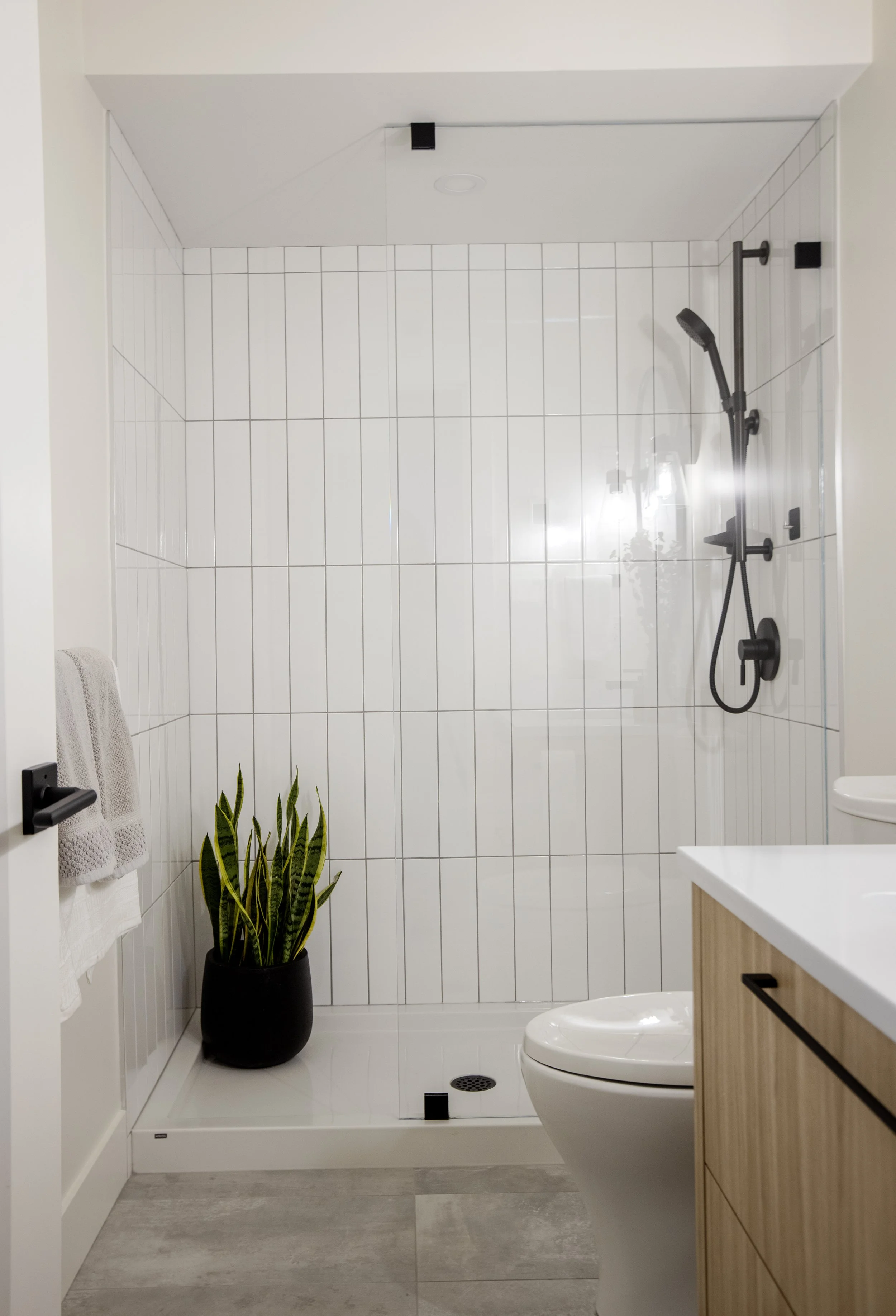 A modern bathroom with a walk-in shower with white tiles, a black shower system, a toilet, a wooden vanity, a plant in a black pot, and white towels hanging on a wall.