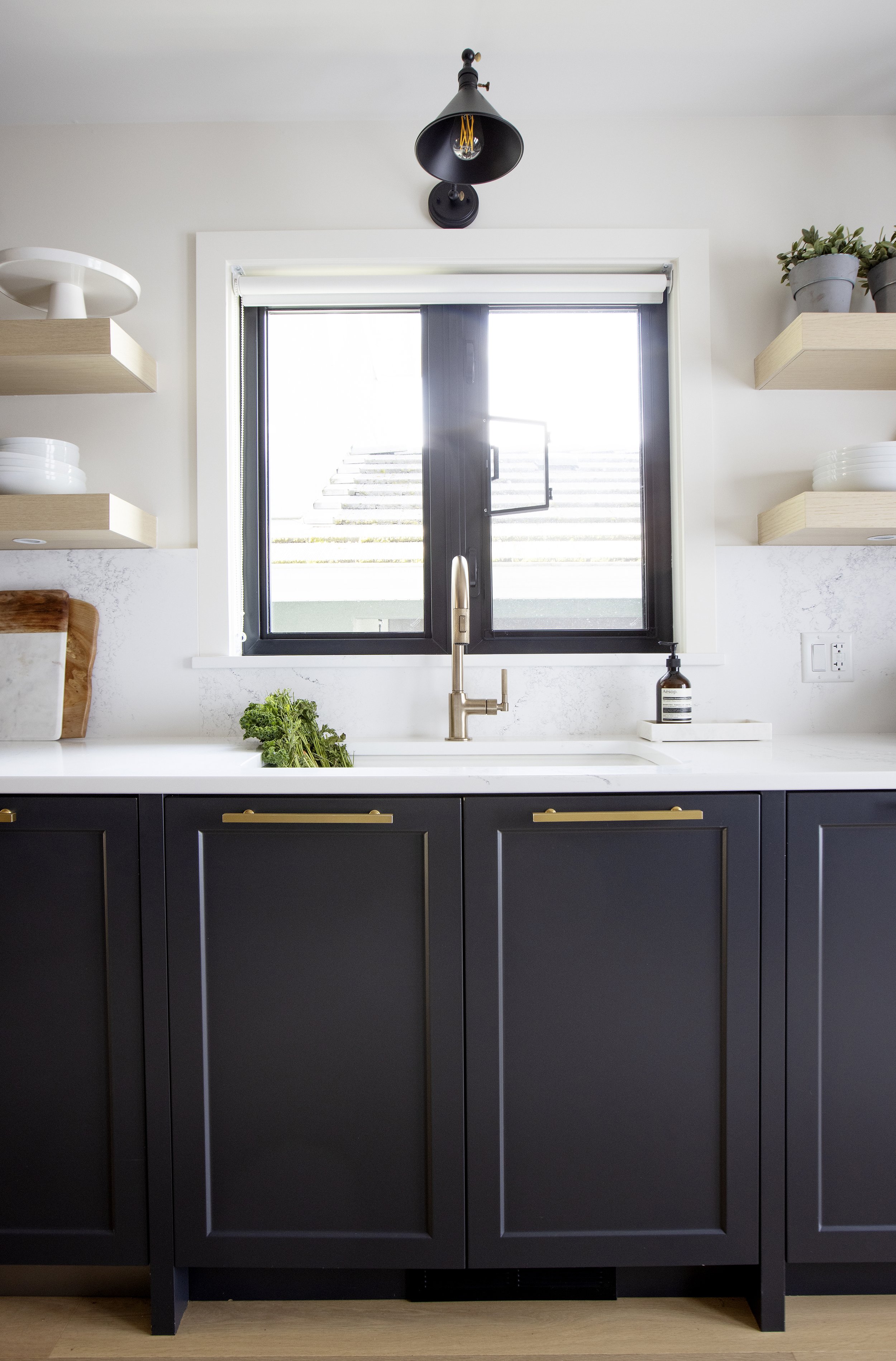 A modern kitchen with dark navy cabinets, white countertop, a window with a view of stairs outside, a gold faucet, a plant, a soap dispenser, and floating shelves with dishes and potted plants.