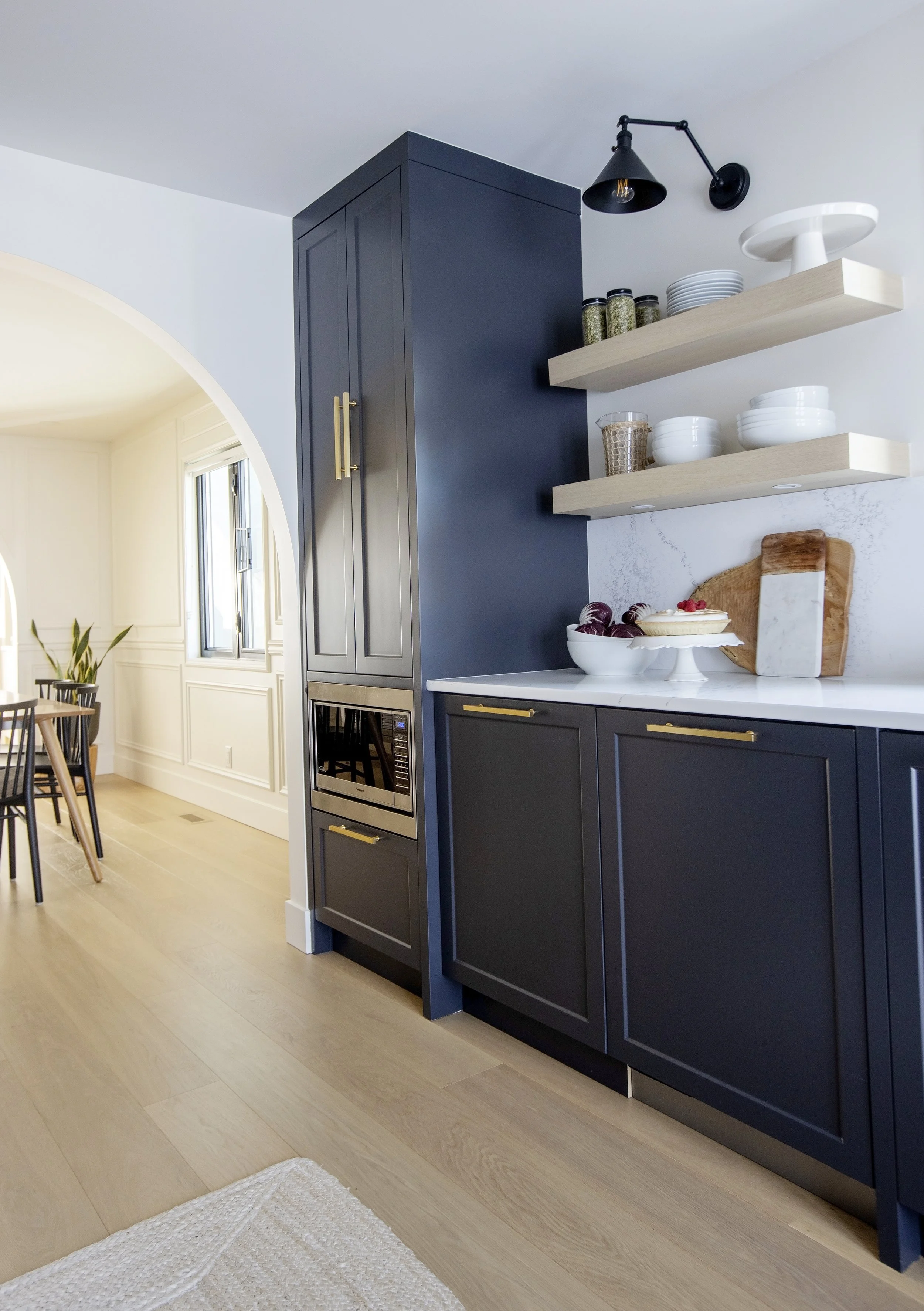 Modern kitchen with navy blue cabinets, open floating shelves with white dishes and jars of herbs, and a black swing-arm wall sconce. Light wood flooring and a white marble countertop.