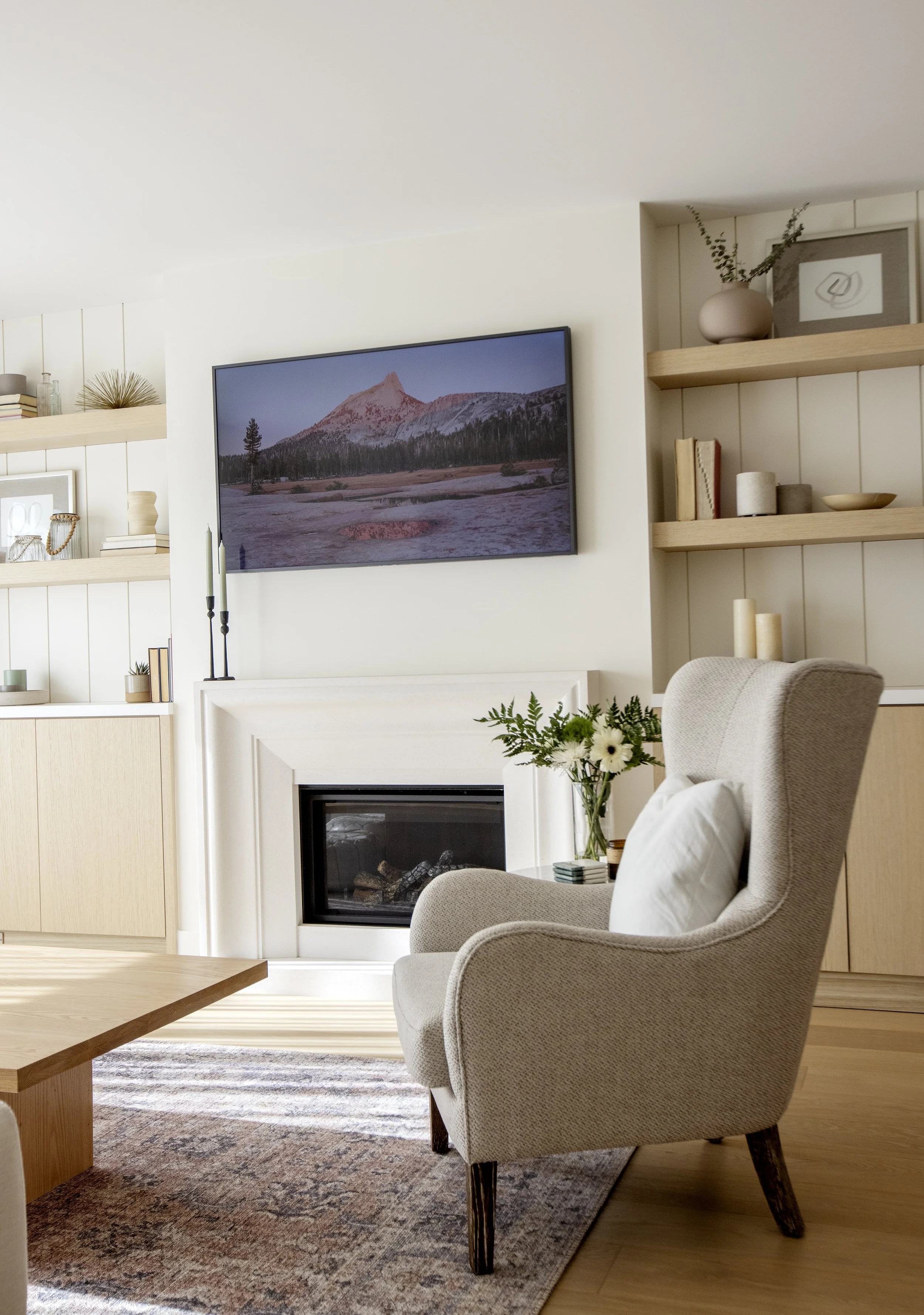 Living room with beige armchair, wooden coffee table, fireplace with white frame, TV on wall displaying mountain landscape, beige built-in shelves with books, vases, framed art, candles, a vase with flowers, rug, and hardwood floors.
