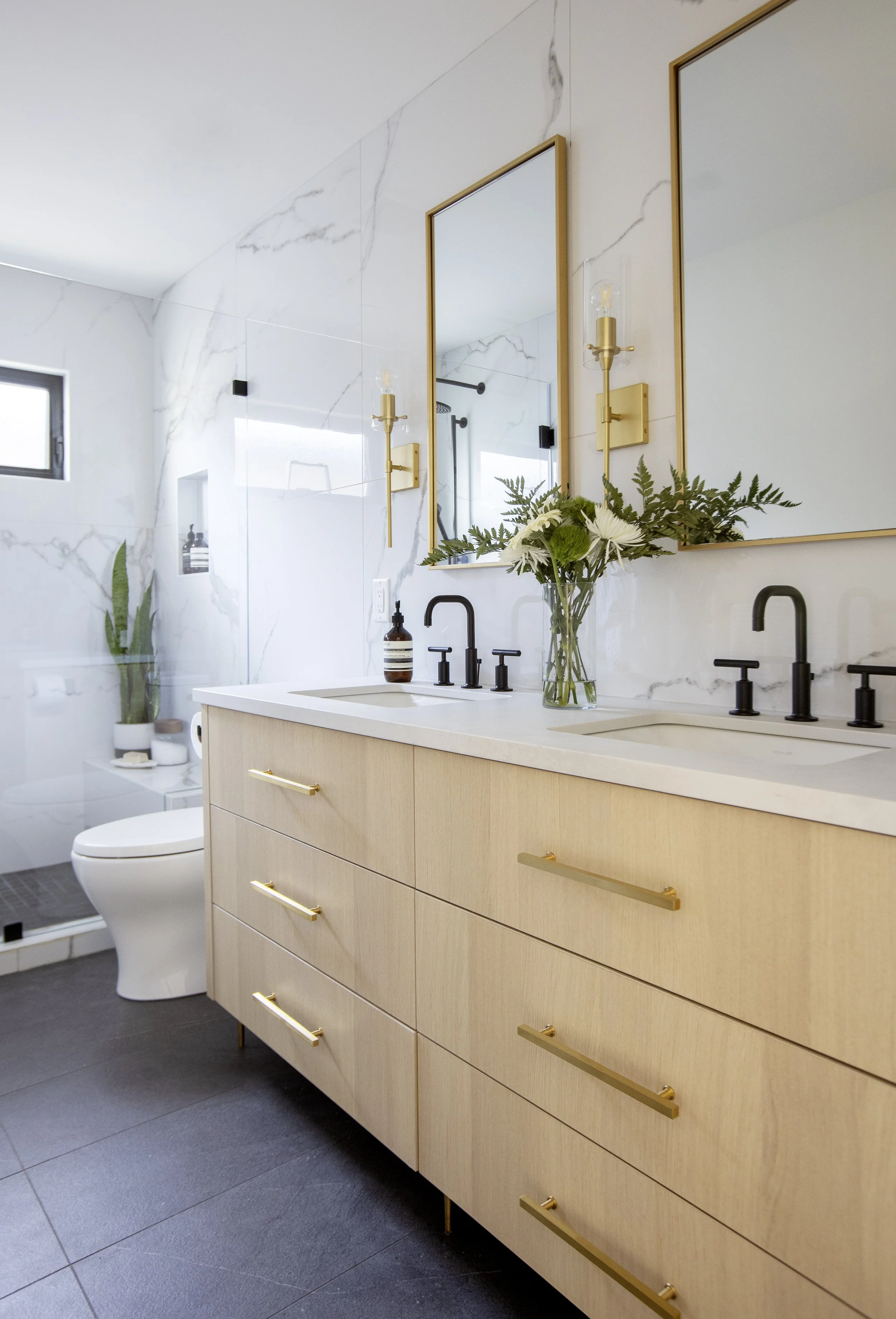 Modern bathroom with a beige wooden vanity, black faucets, a mirror with gold frame, white marble wall tiles, a small window, and a vase with white flowers.