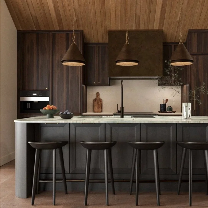 Modern kitchen with dark wood cabinets, a white marble island with seating, pendant lights, and minimal decor including a fruit bowl and a vase with greenery.