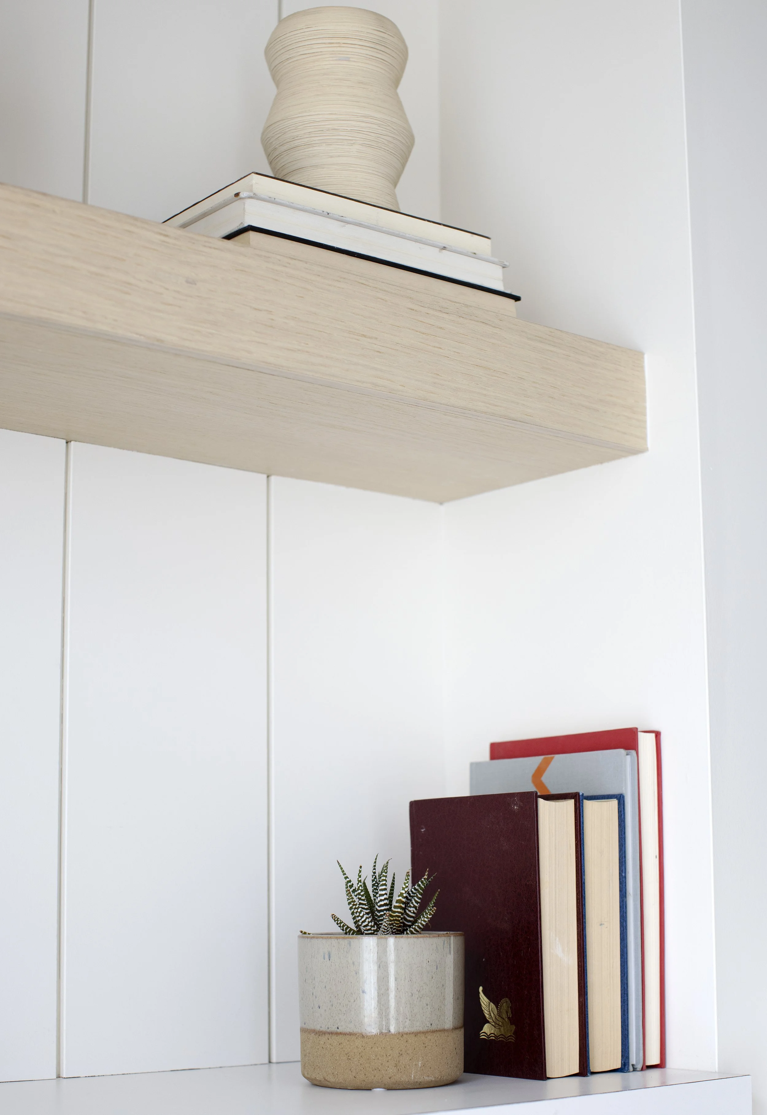 A white shelf with a wave-shaped beige ceramic vase on top, and a small potted plant and several books with colorful spines underneath.