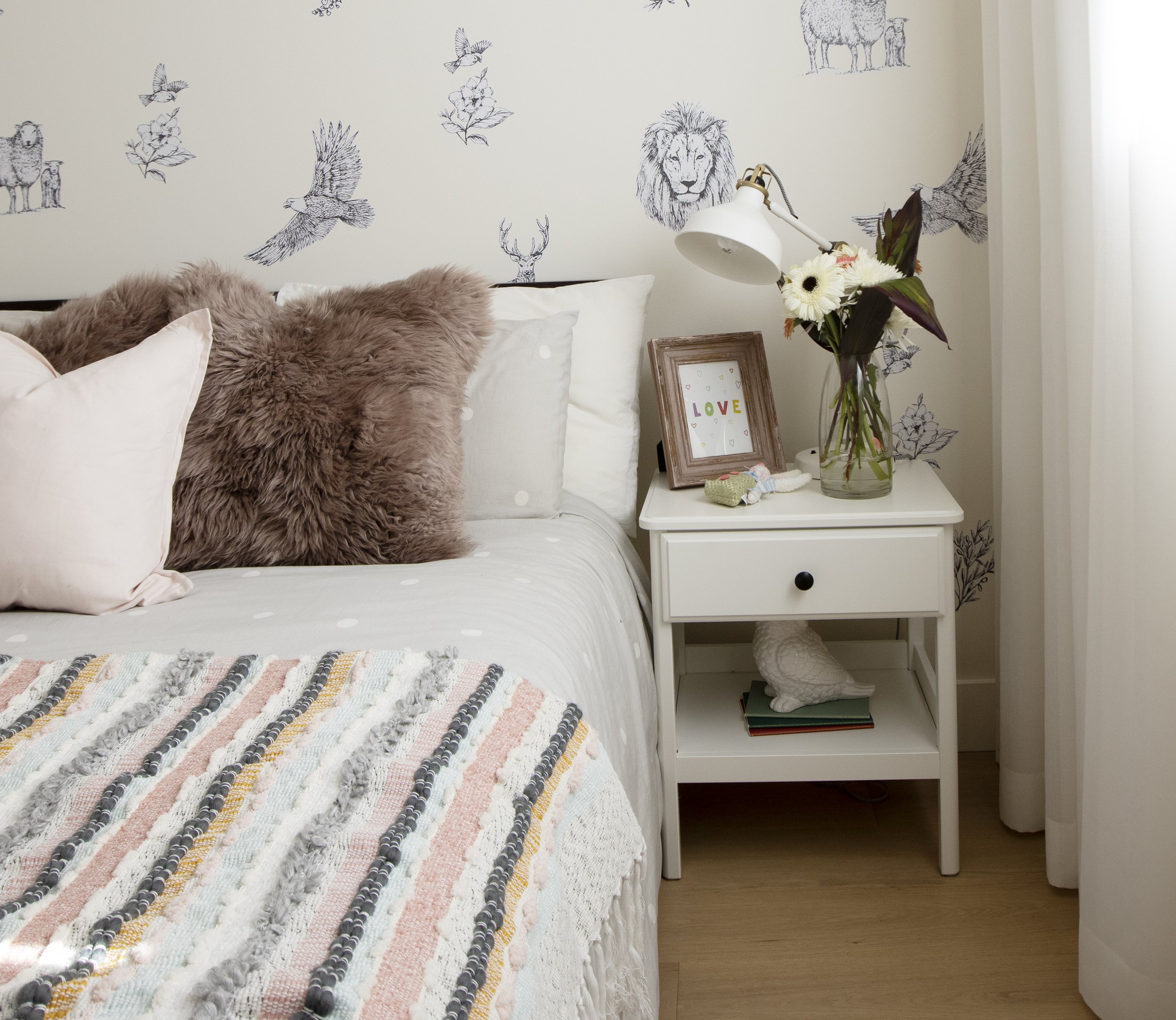 View of a bedroom corner with a bed, decorative pillows including a furry pink one, a bedside table with a flower vase, a framed picture, a lamp, and wall art featuring animal sketches and birds.