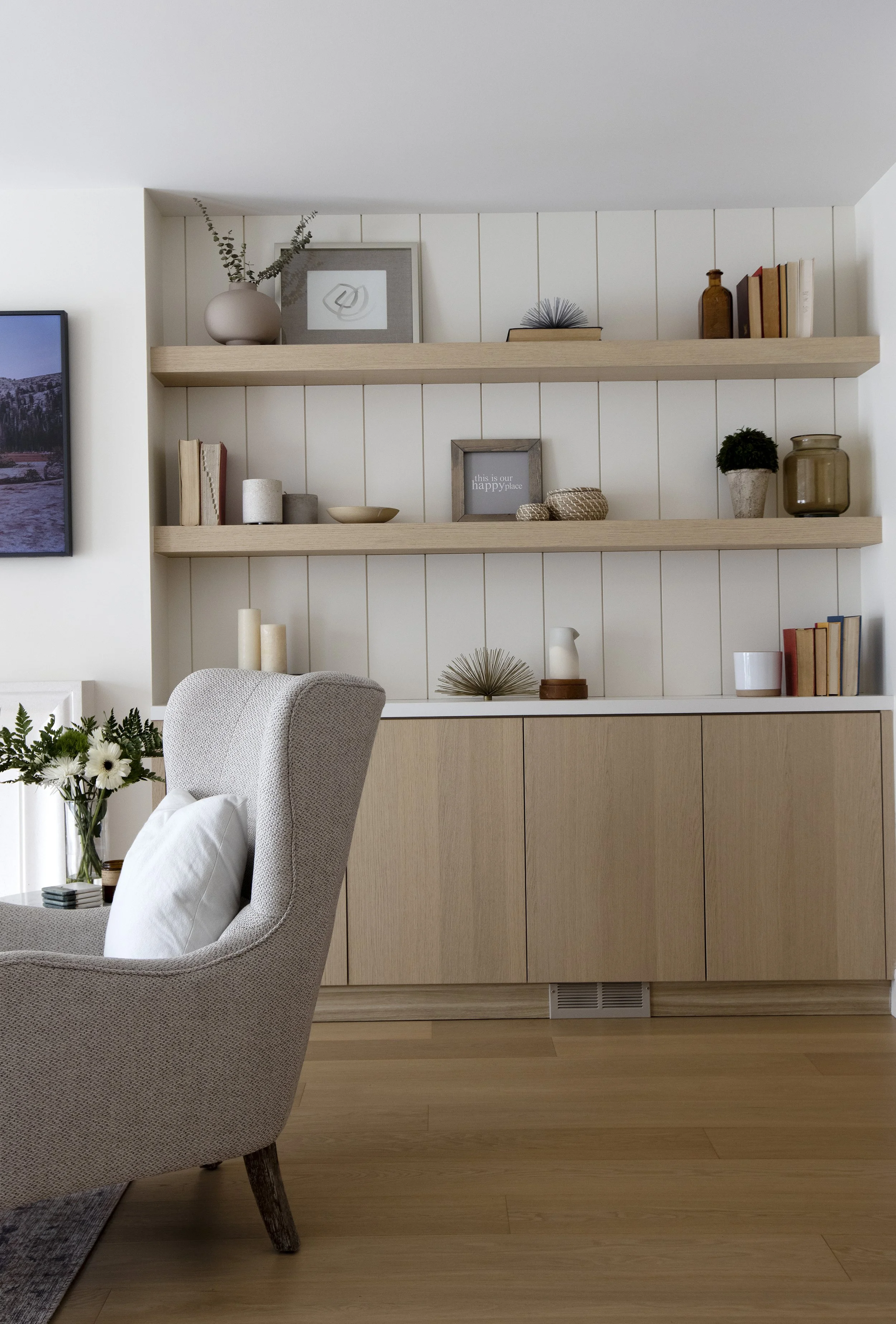 A cozy living room with a beige armchair, a white pillow, a glass vase of white flowers, a gray rug, and light wooden flooring. Behind the armchair is a built-in cabinet with white paneling and two open wooden shelves above, decorated with books, fra