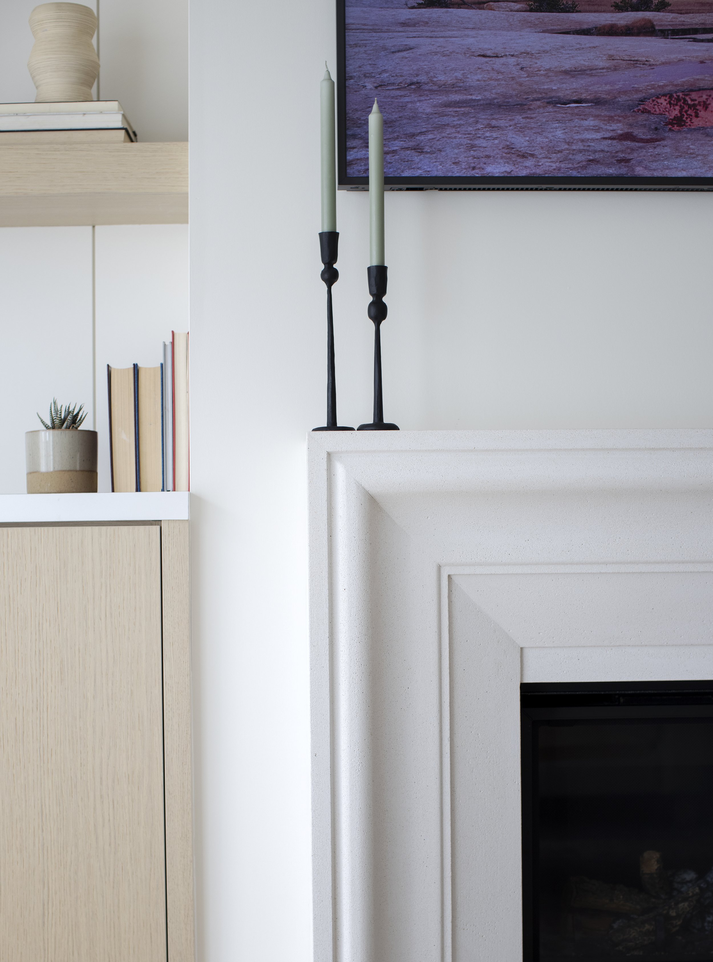 A close-up of a modern white fireplace with two tall black candlesticks with unlit green candles on top, positioned on the mantel. To the left, there is a light wooden cabinet with books and a small plant in a beige pot. Part of a wall-mounted TV dis