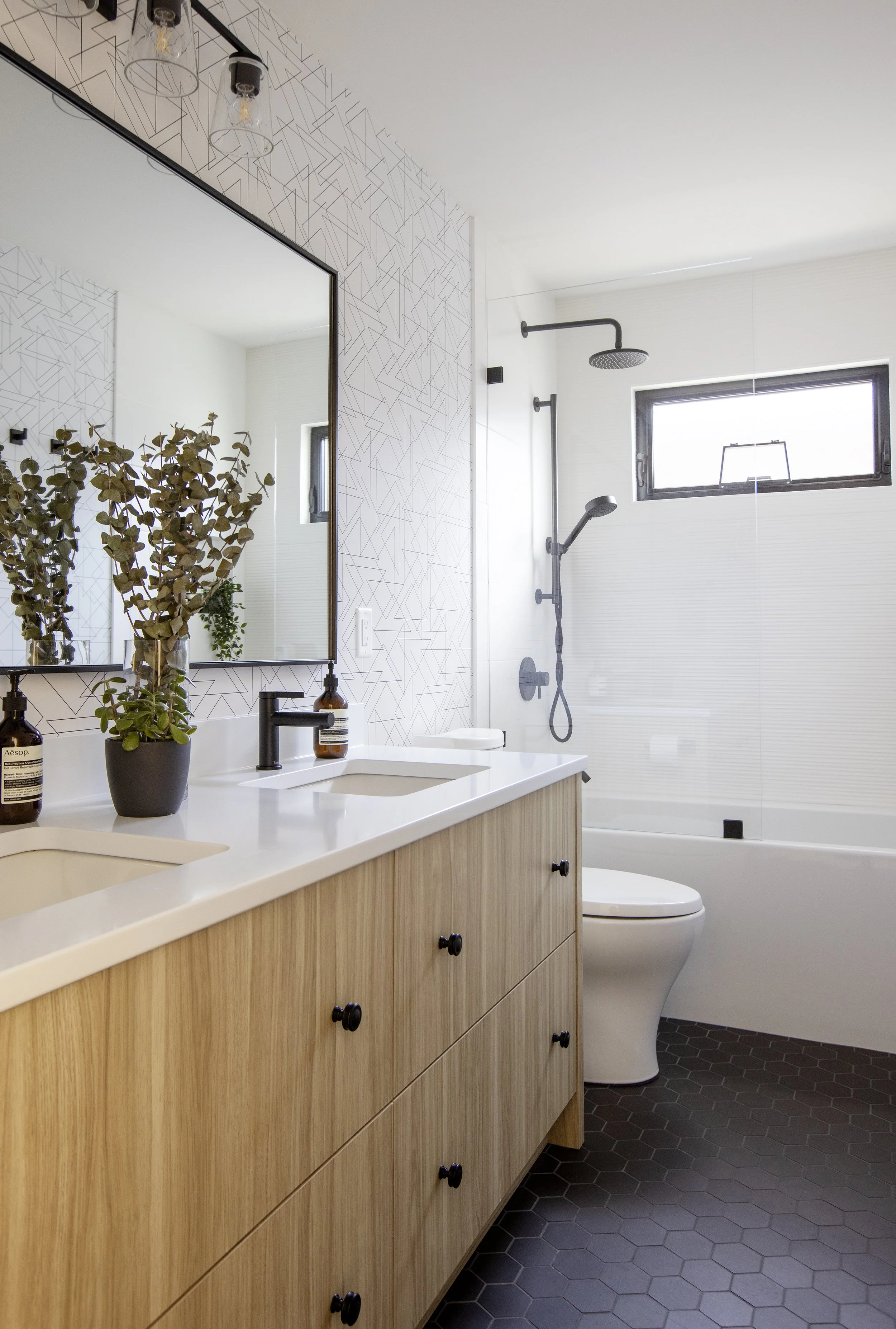 Modern bathroom with light wood vanity, white countertop, plants, large mirror, geometric patterned wall, black fixtures, shower area with black showerhead and window, dark hexagonal floor tiles.
