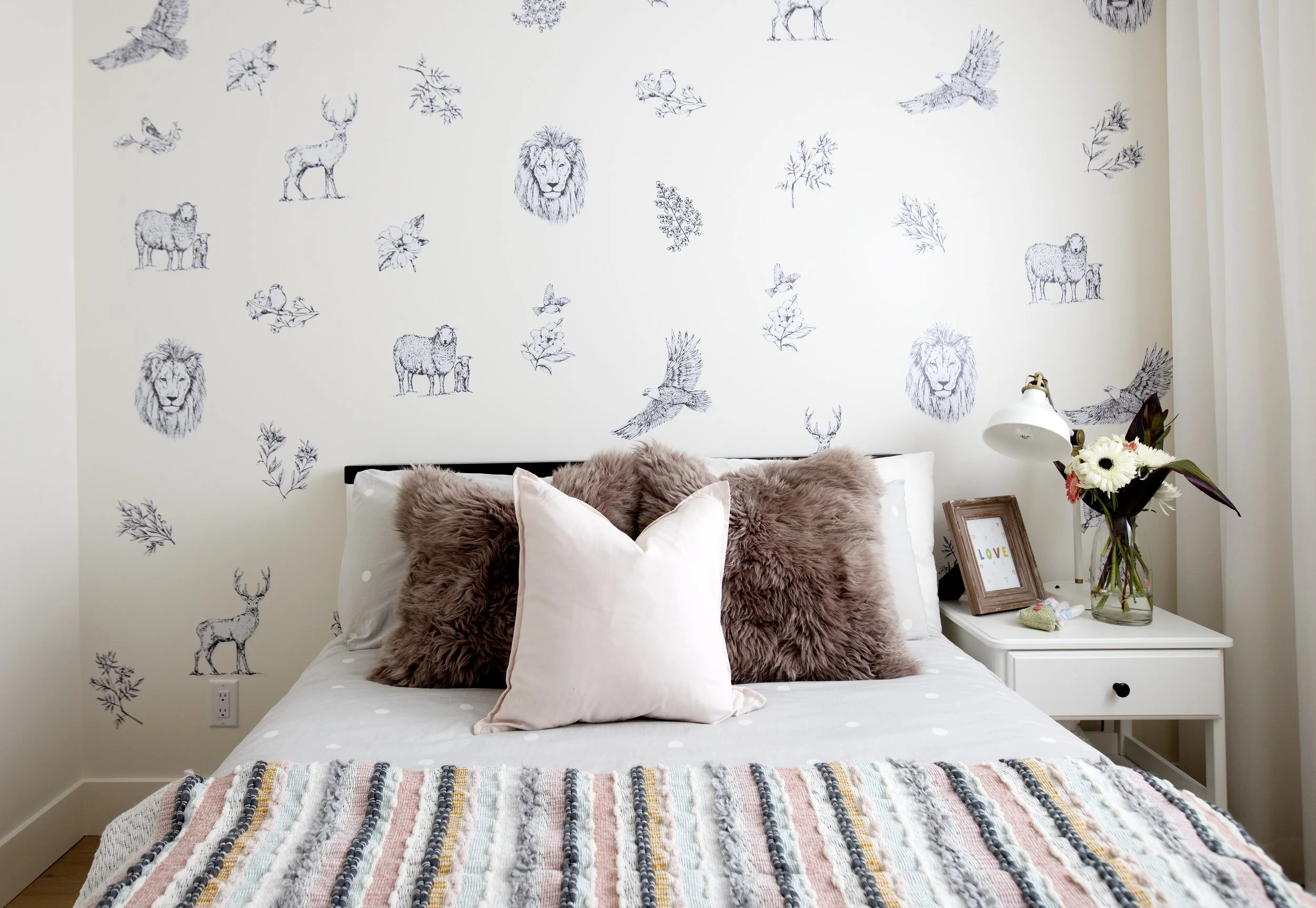 A bedroom with a white wall decorated with black line drawings of various animals, including lions, deer, birds, and rabbits, along with some botanical illustrations. The bed has a multicolored striped throw blanket, a fluffy brown pillow, and a ligh