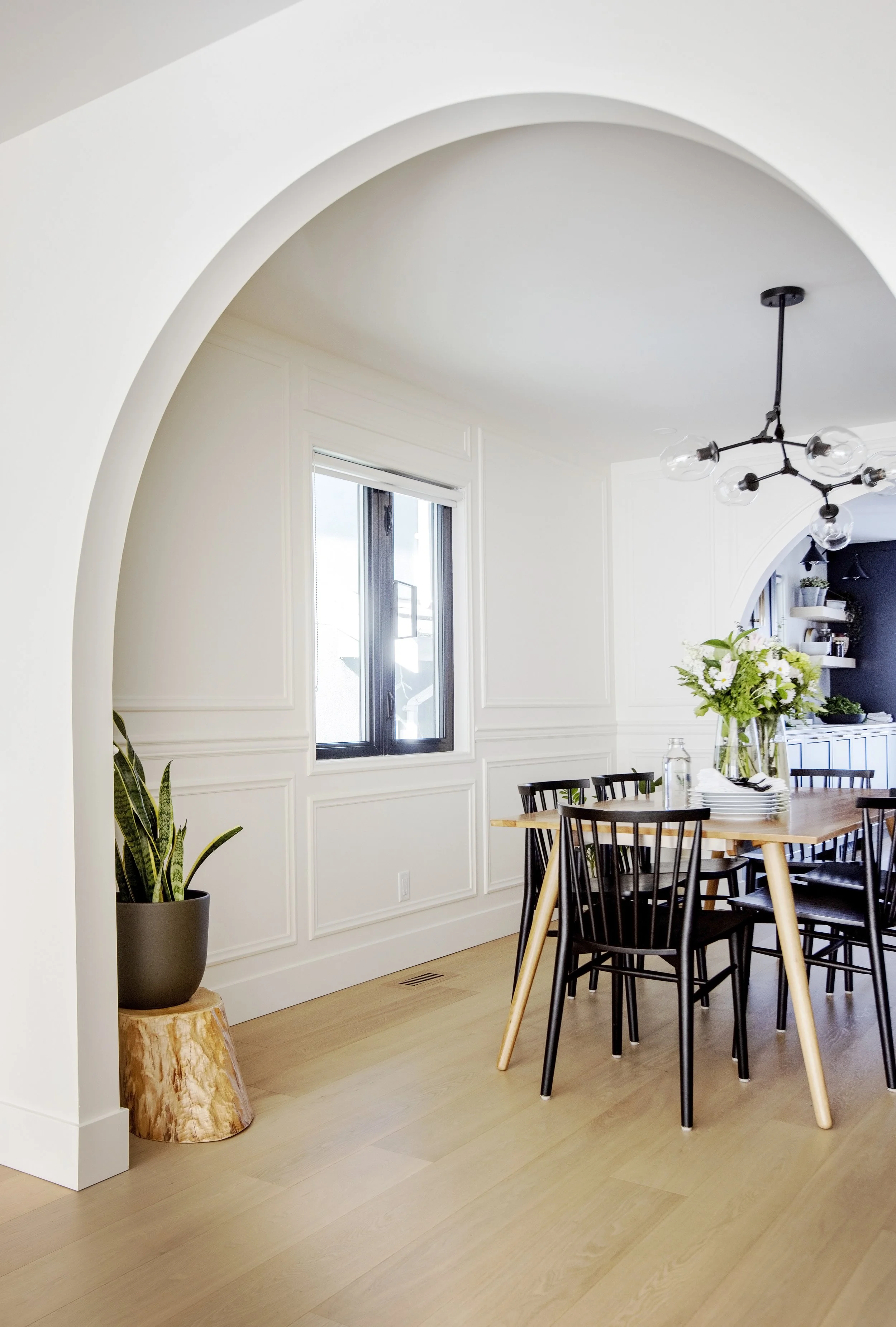 A dining room with a wooden table, black chairs, a glass vase with white flowers, a large window, and a modern black chandelier.