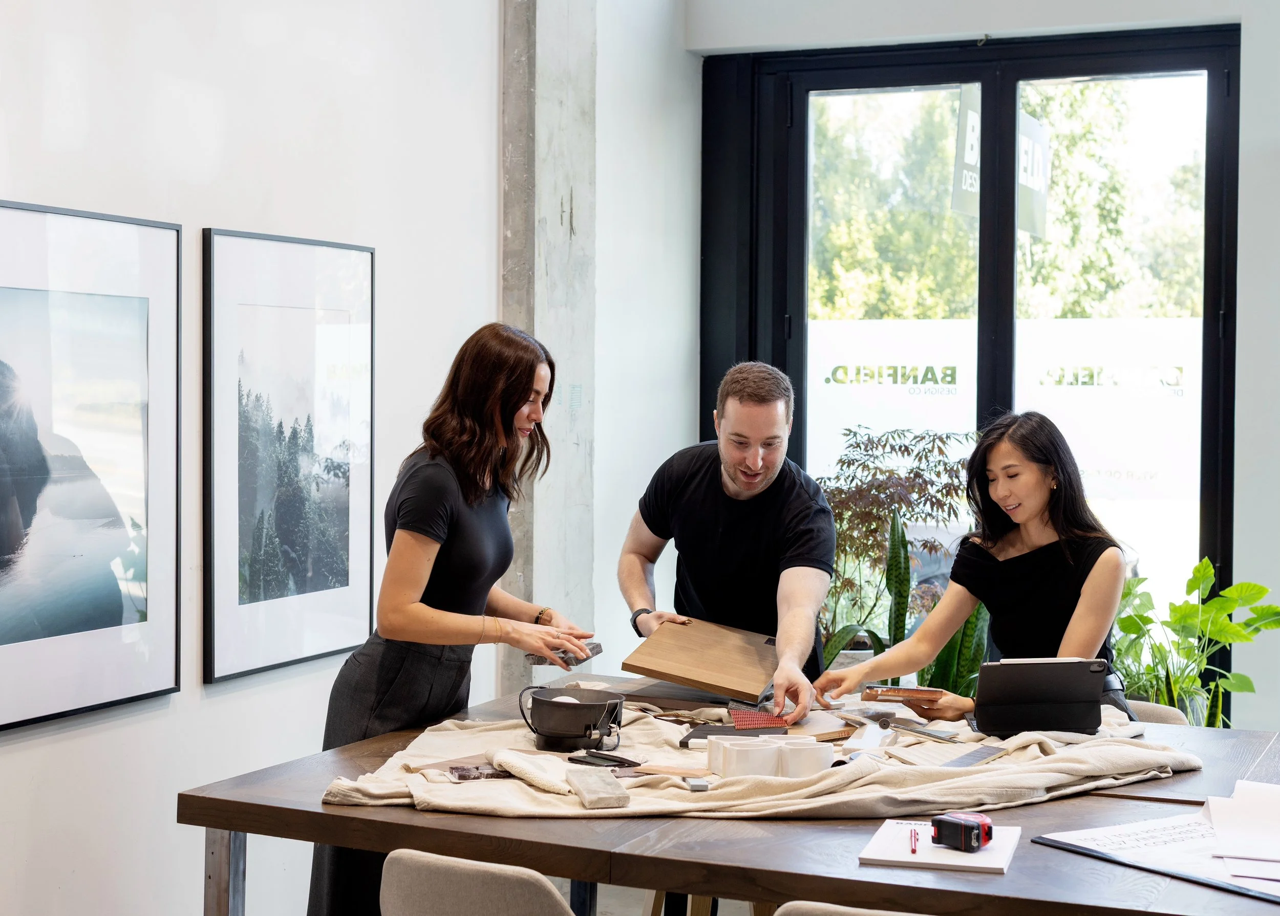 Three people working at a table with various fabric samples and tools, in a well-lit modern room with large windows and framed artwork on the wall.