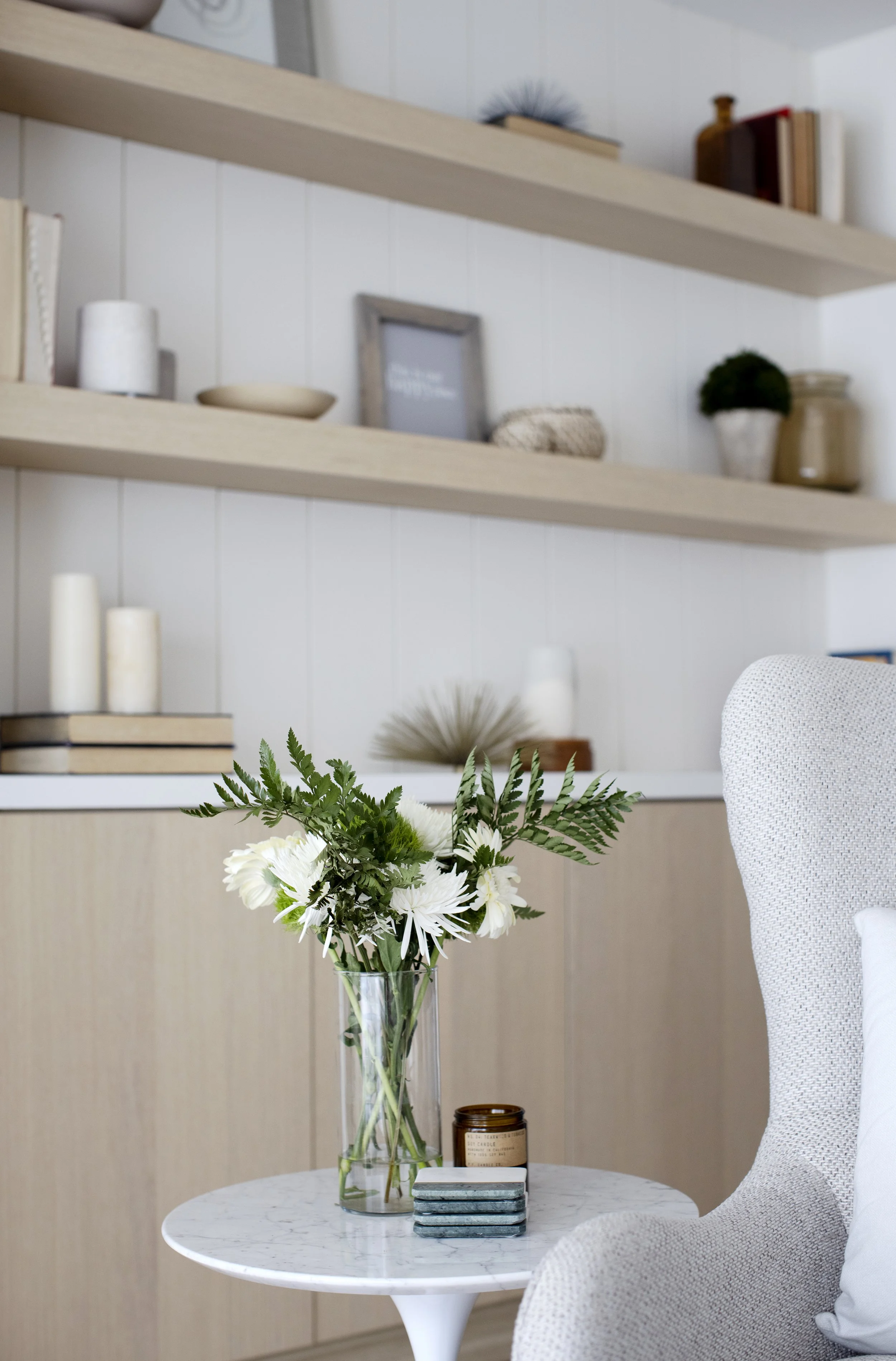 A white marble round table with a glass vase containing white flowers and green foliage, a brown candle, and a phrasebook or small books stacked on top of each other, in a modern, bright living space with beige and white decor and shelves in the back