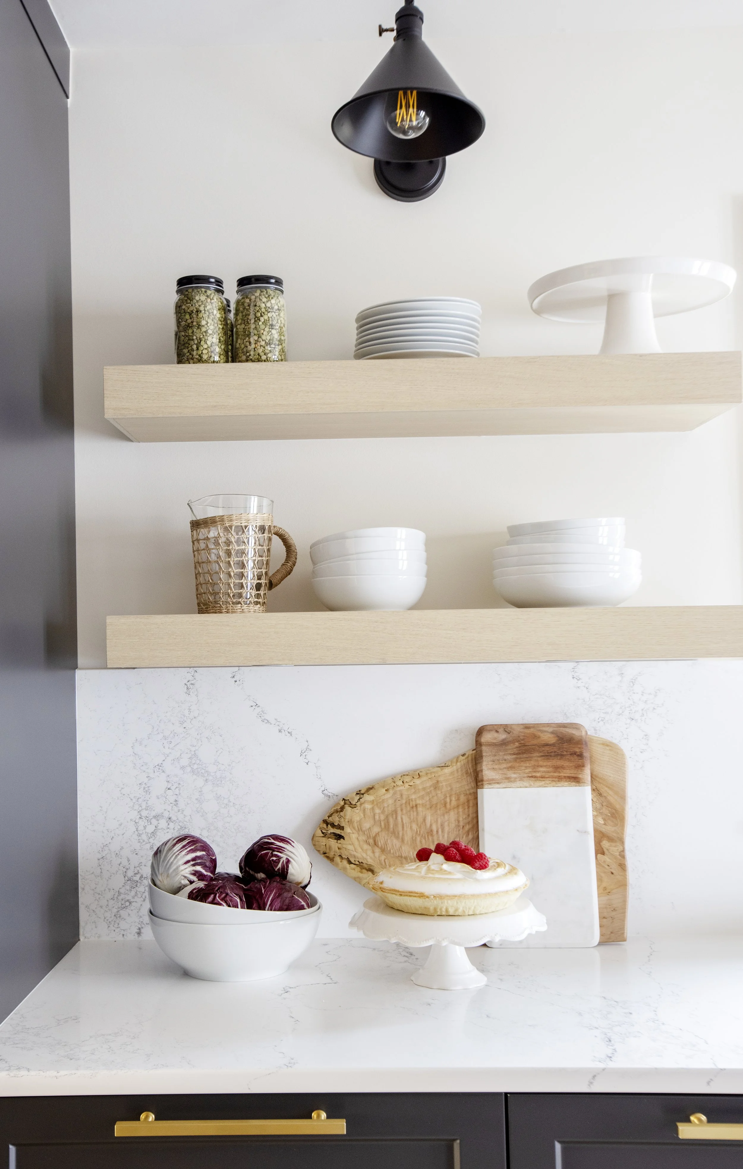 Open kitchen shelves with jars of grains, white plates, and a white cake stand. Below, a marble countertop with a bowl of radicchio, a cake with raspberries, and an cutting board are visible.