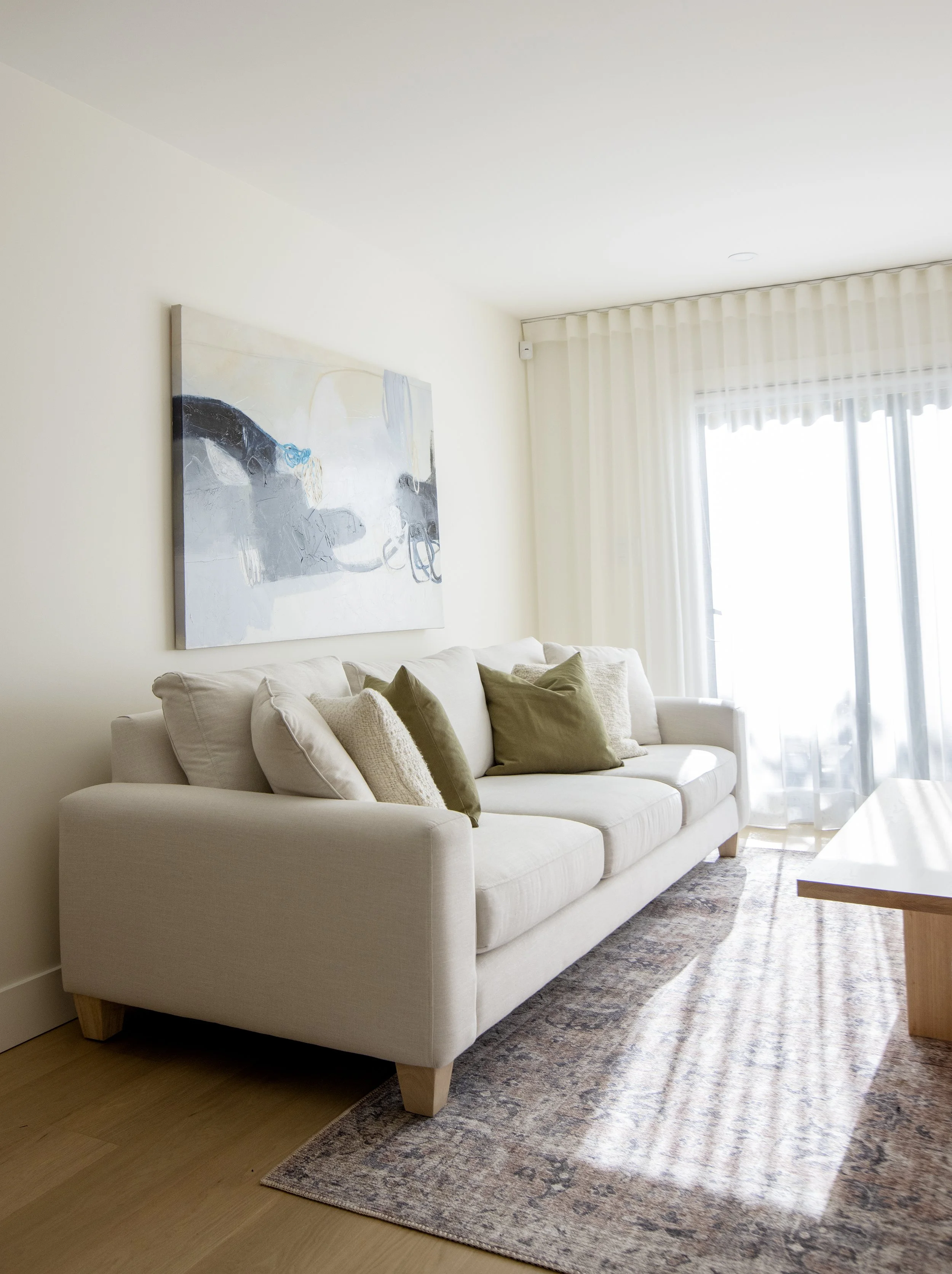 A bright living room with a white sofa, beige and green pillows, a wooden coffee table, patterned rug, large window with sheer curtains, and an abstract painting on the wall.
