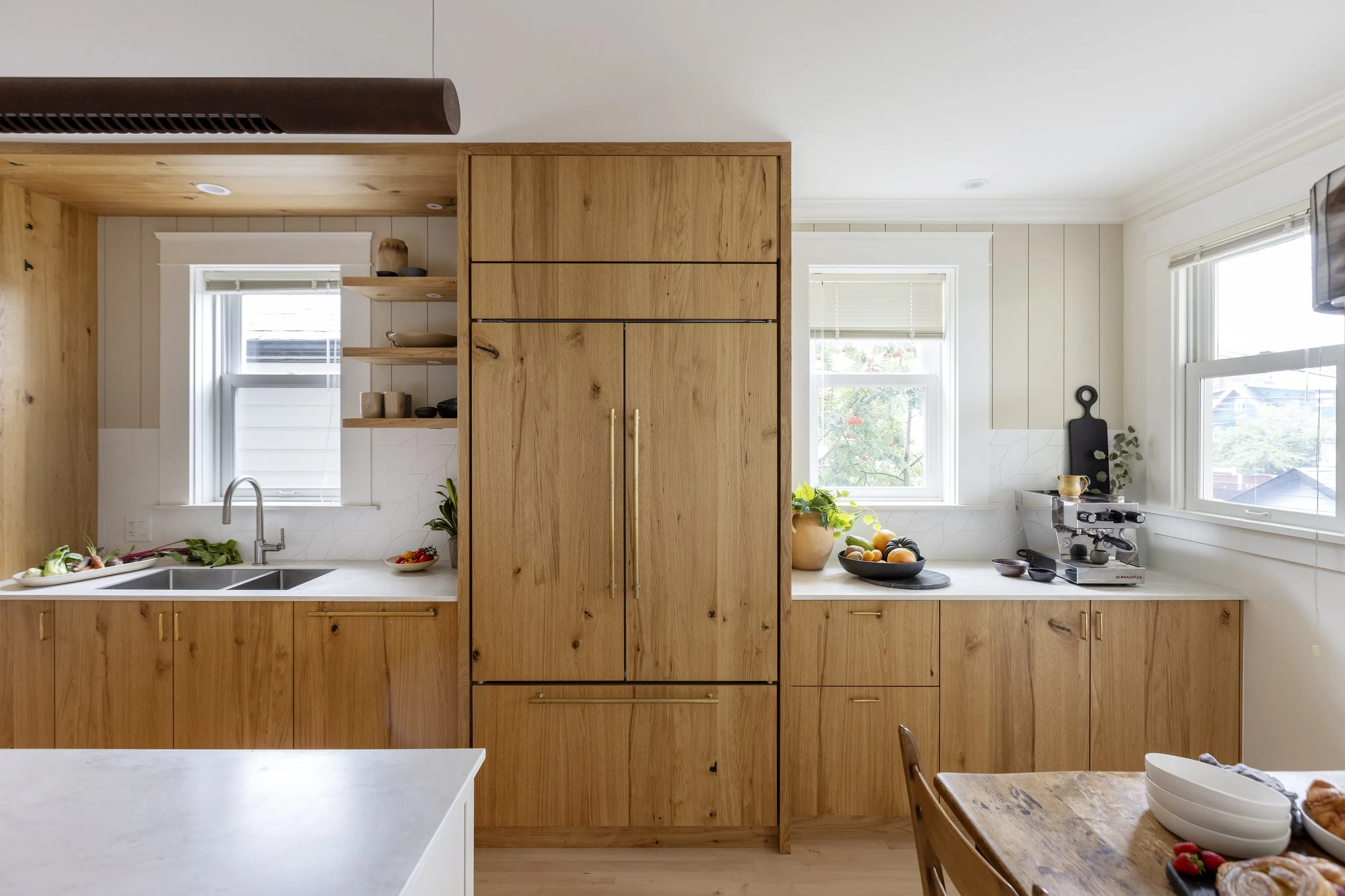 Modern kitchen with natural wood cabinetry, white countertops, and two large windows letting in natural light. Decor includes bowls of produce, a coffee machine, and decorative items.